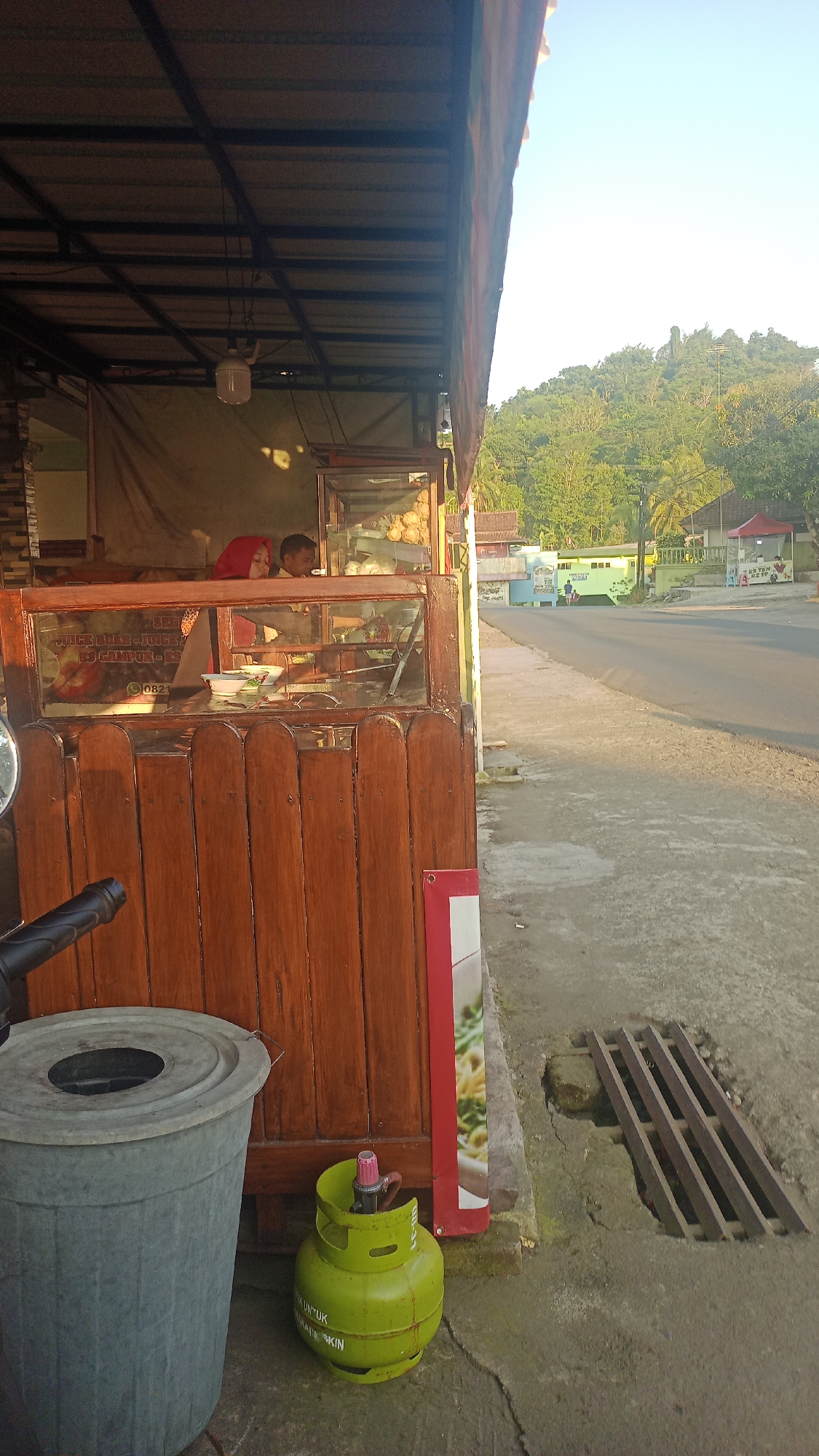 The image depicts an outdoor food stall in Karyamandala, Indonesia, with a road and a lush green hillside in the background. The stall features a wooden counter with a glass display case, showcasing various food items. Two individuals, possibly vendors, are visible behind the counter. In the foreground, a large grey trash bin and a bright green gas cylinder are placed on the ground, adjacent to a wooden structure. A metal grate covers a drainage channel on the paved ground. The scene is bathed in the warm light of what appears to be either early morning or late afternoon.