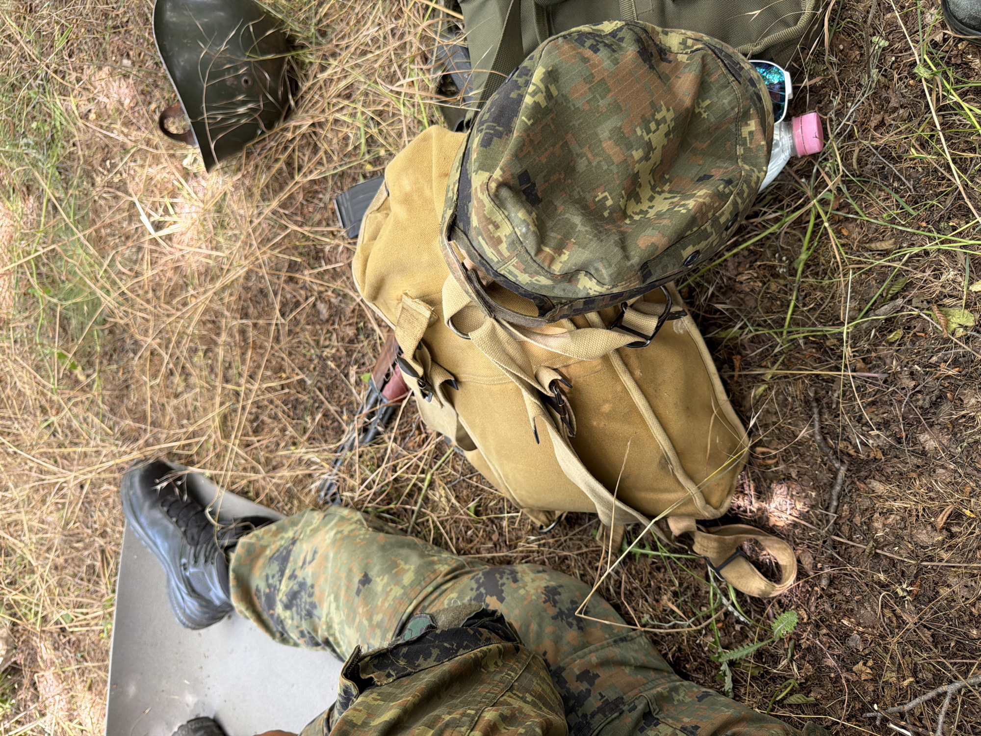 A high-angle, medium shot captures military equipment resting on dry grass and dirt. The scene is set outdoors, likely in a natural environment. In the foreground, a person's leg clad in camouflage fatigues is visible, with a black combat boot resting on a grey mat. Adjacent to the leg are two bags: one a light tan canvas backpack with straps, and another bag, possibly a helmet cover, in camouflage pattern. A green helmet with a brown strap is positioned to the right of the bags. A pink-capped water bottle and sunglasses are partially visible underneath the camouflage bag. In the background, dry grass and sparse vegetation cover the ground. The image does not provide enough information to determine the specific location within Beliakovets, Bulgaria.