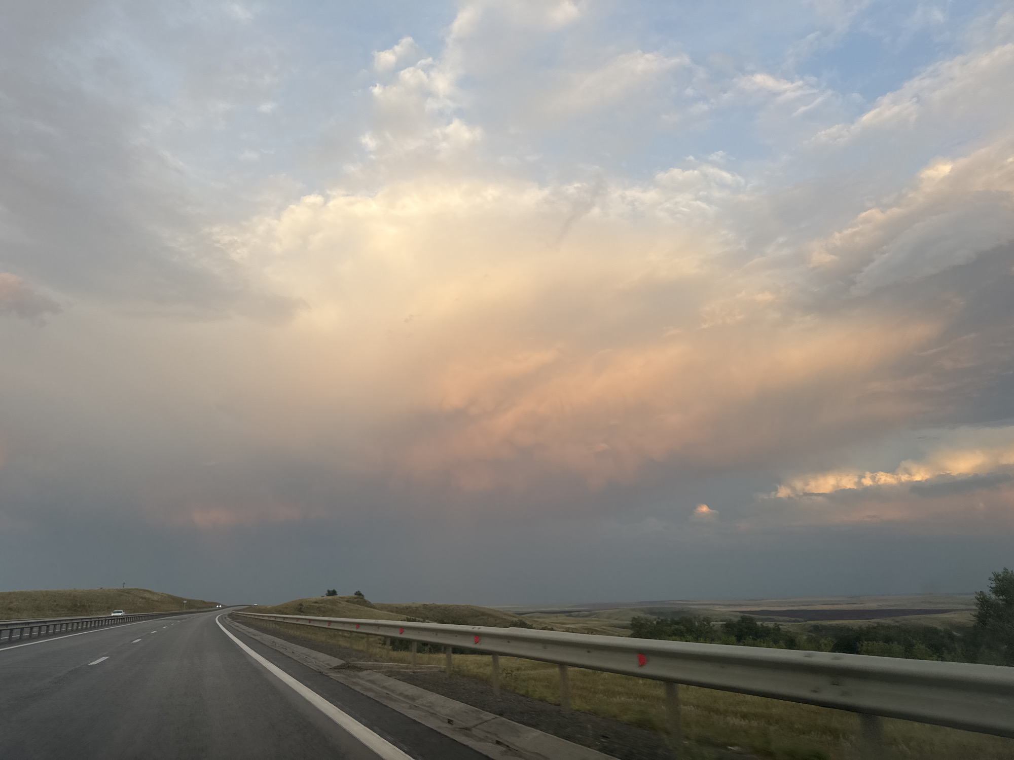 The image shows a highway in Syrt, Russia, during sunset. The sky is filled with dramatic clouds, colored in shades of orange, pink, and gray, with patches of blue visible. The road, a multi-lane highway, curves to the left and disappears into the distance. A metal guardrail runs along the right side of the road, with red reflective markers. The landscape on either side of the highway is characterized by rolling hills covered in dry grass, with some scattered trees. In the distance, a few vehicles can be seen on the road. The overall impression is one of a journey through a vast and open landscape under a breathtaking sky.