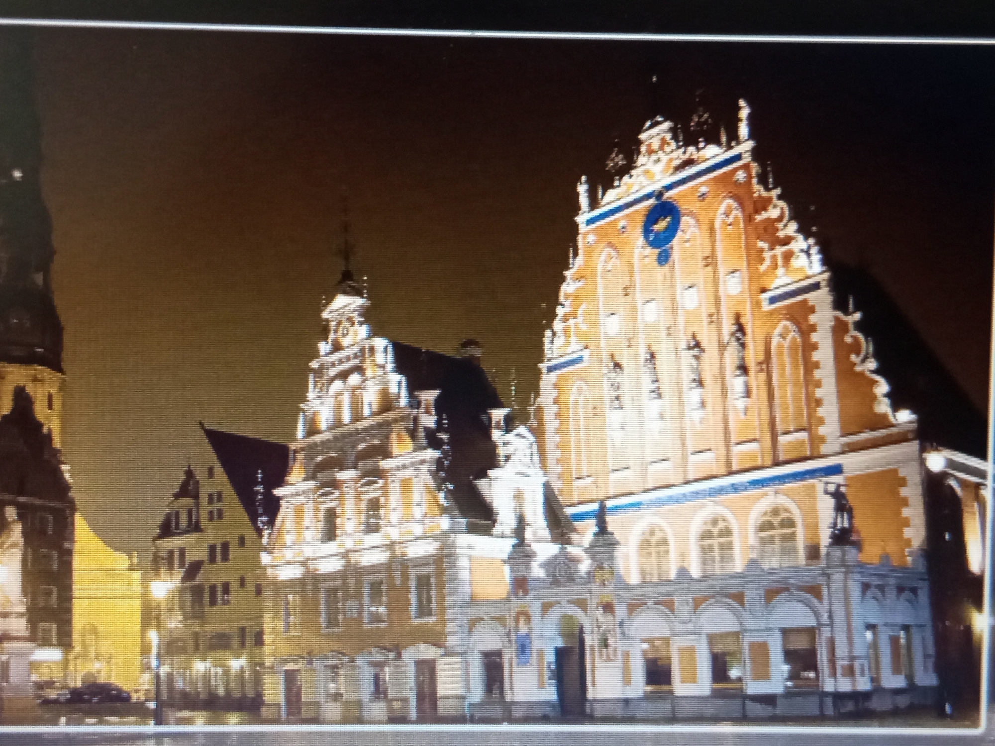 The image shows a nighttime view of the House of the Blackheads and the surrounding buildings in Riga, Latvia. The architecture is ornate and features brightly lit facades, with the House of the Blackheads being the most prominent structure. It is a large, reddish-brown building with white decorative elements and a steep, stepped gable. Several statues are visible on the building's exterior, and a blue circular emblem is mounted high on its facade. Adjacent to it, to the left, is another building with a similar style, though less elaborate, also illuminated. Further to the left and in the background, other buildings are visible, some with pointed roofs characteristic of old European cities. The scene is illuminated by artificial lights, suggesting it is after dark. The sky is dark and appears clear. No people are clearly discernible in the image, though there might be shadows or distant figures that are not distinct. The overall impression is one of historical grandeur and a vibrant urban setting at night.