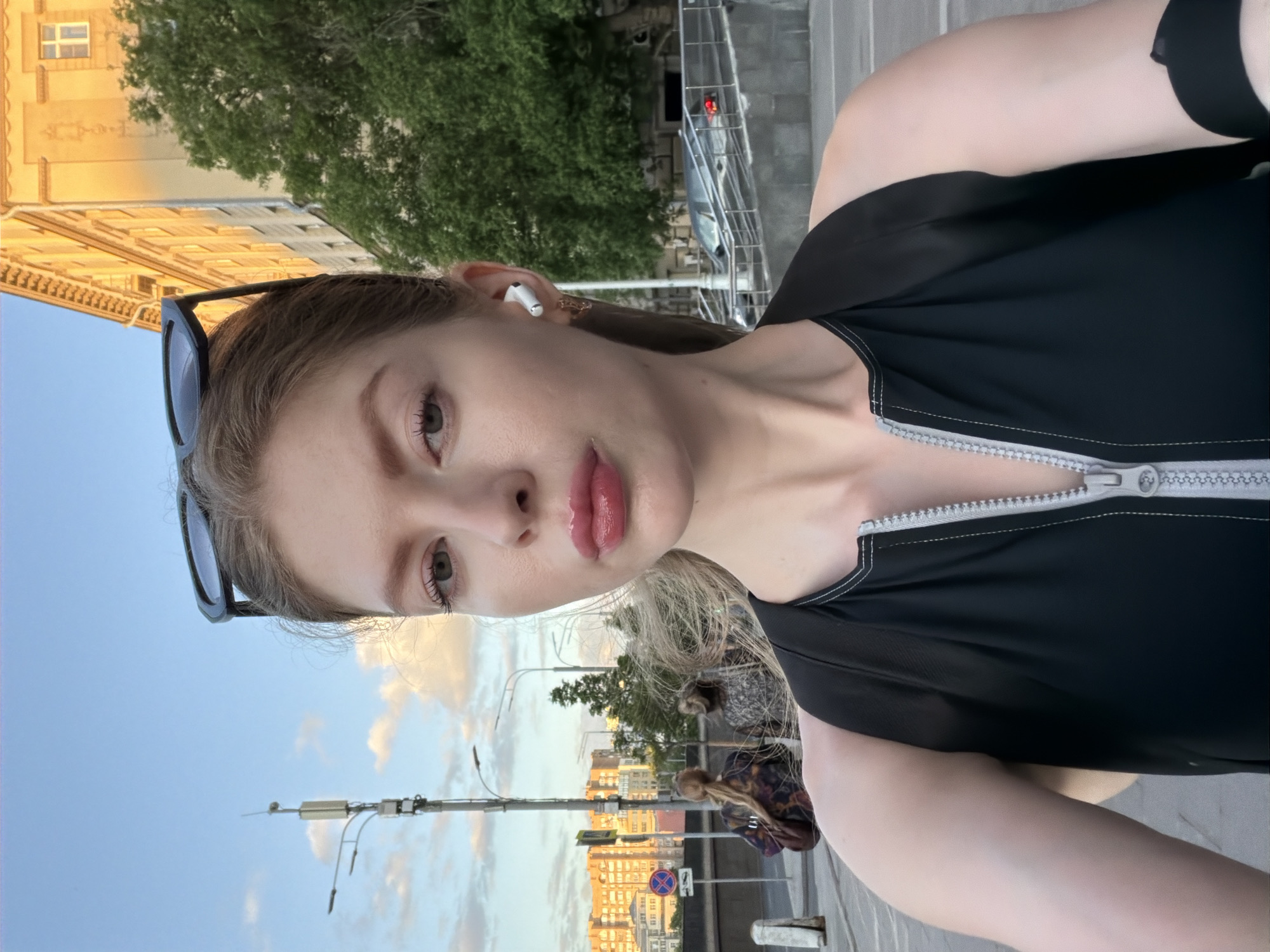 A woman takes a selfie. She has fair skin, blond hair, and light eyes. Sunglasses are perched on her head. She is wearing an earbud in her left ear. She is wearing a black zip-up top.
She is outside in an urban setting, likely in Moscow, Russia. In the background is a street with buildings, trees, street lights, and pedestrian crossing signs. The buildings are a light orange color. There are people walking on the sidewalk. The sky is light blue with some clouds.