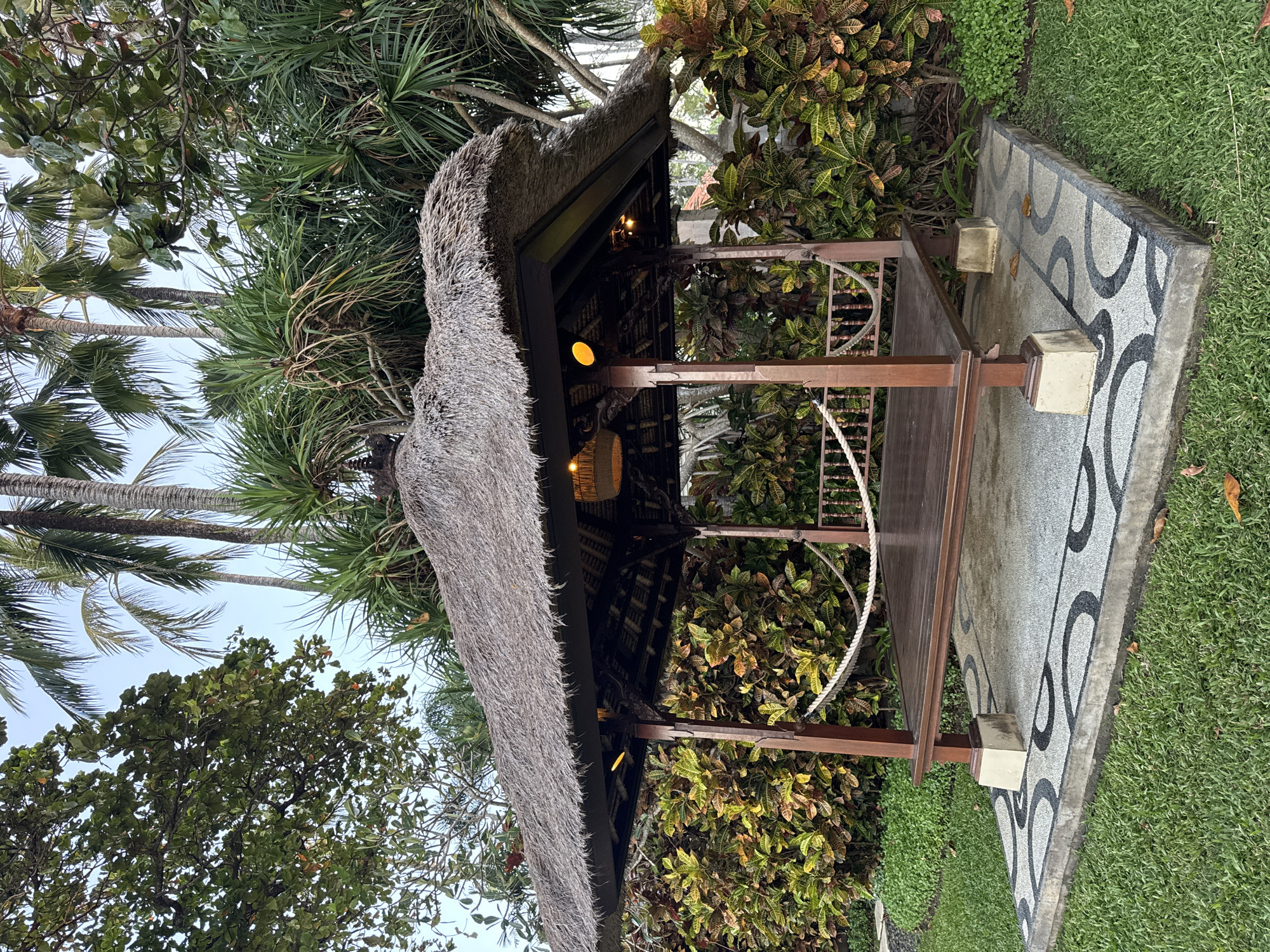 An outdoor, open-sided, wooden pavilion with a thatched roof stands on a paved square surrounded by lush greenery and palm trees. The pavilion's floor is made of dark wood, and a thick rope railing runs along the sides. There are two hanging lights inside the pavilion. The paved square features a decorative mosaic pattern of black and gray circles. The surrounding area is covered in neatly trimmed green grass, with dense bushes and foliage behind the pavilion. The sky above is a clear, pale blue. The scene appears to be a tranquil outdoor setting, possibly within a resort or garden in Kuta Selatan, Indonesia.