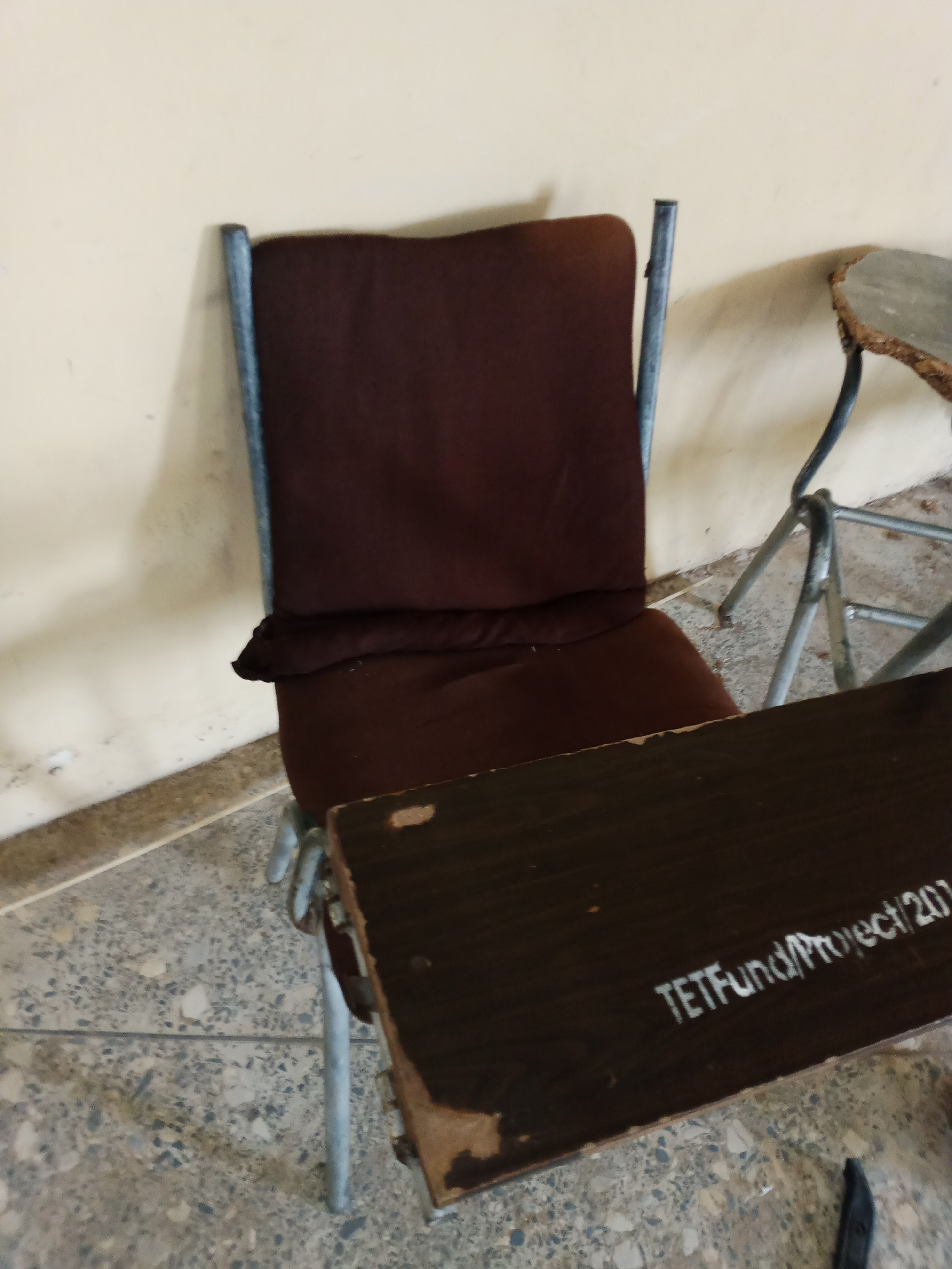 The image shows a close-up of a brown cushioned chair with a wooden desk in front of it. The chair appears to be in a classroom or similar institutional setting, possibly in Jalingo, Nigeria. The desk has "TETFund/Project 20" written in white paint on its surface. The wall behind the chair is a light beige color, and the floor is a terrazzo-like material. Another chair or table leg is visible to the right of the frame. The scene appears to be poorly lit and the objects are somewhat aged, showing signs of wear and tear. There are no people visible in the image.