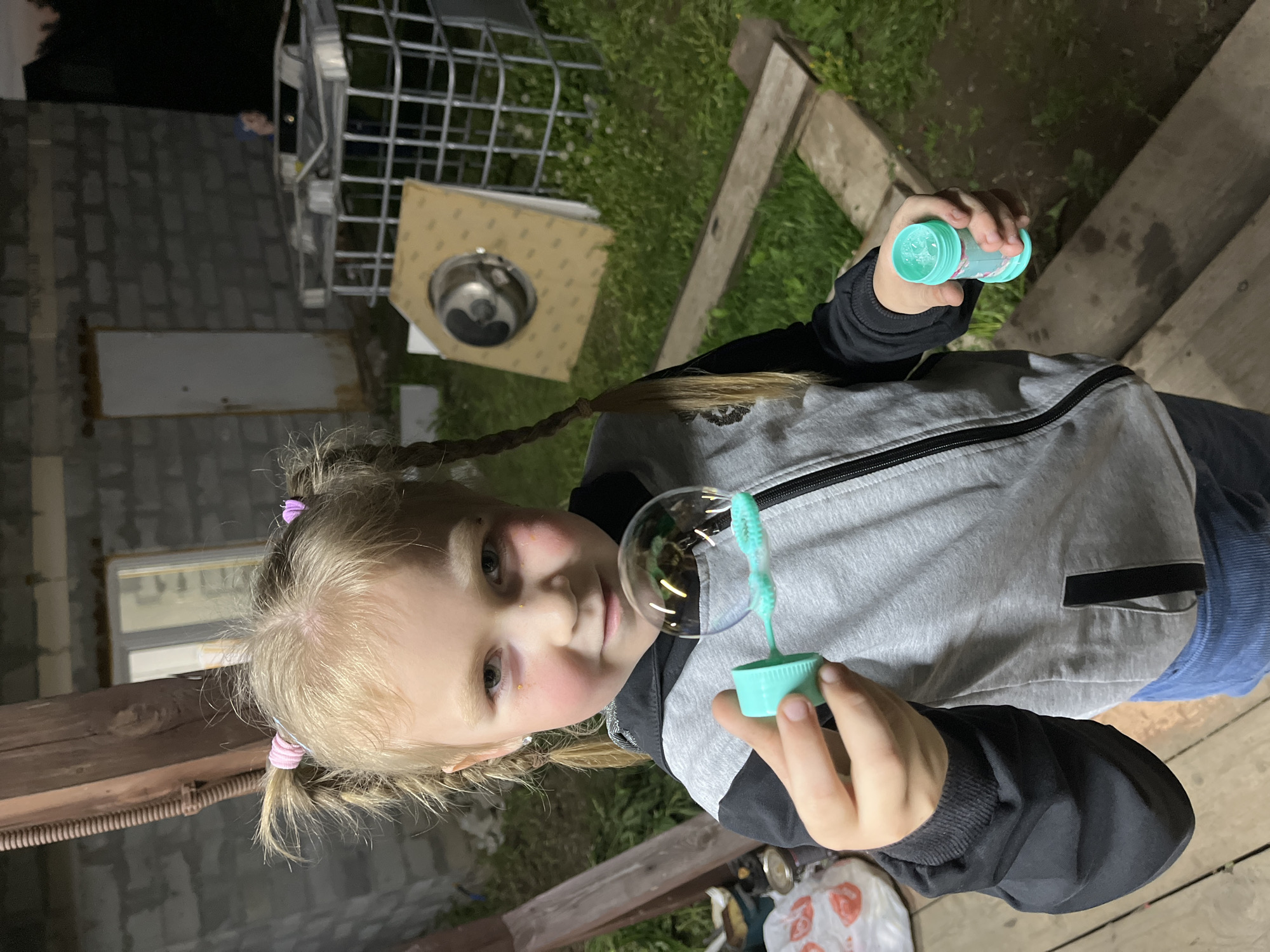 A young blonde girl with pigtails stands outdoors, holding a bubble wand and a container of bubble solution. She is looking at the camera. She wears a gray jacket with black accents. A large bubble floats in front of her face. The setting appears to be a backyard or garden area. In the background, there is an IBC container, a gray-walled structure, and vegetation. Discarded trash in the foreground. There is no information in the image to confirm this location as Chaikovsky, Russia.