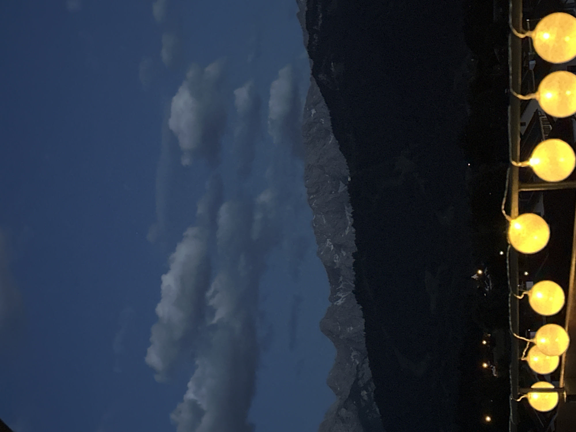 The image depicts a twilight scene overlooking a mountainous landscape. In the foreground, a string of illuminated spherical lights hangs horizontally, casting a warm glow. These lights are attached to a railing, suggesting an outdoor vantage point, possibly a balcony or terrace. Beyond the railing, a dark, tree-covered hillside stretches across the mid-ground. Further back, majestic mountains rise, their peaks dusted with snow and illuminated by the fading daylight. The sky above is a deep blue, streaked with dramatic, dark clouds. In the distance, faint lights of a settlement can be seen nestled among the trees at the base of the mountains, indicating the presence of the town of Garmisch-Partenkirchen. There are no people or animals visible in the frame. The overall impression is one of natural beauty and tranquility.
