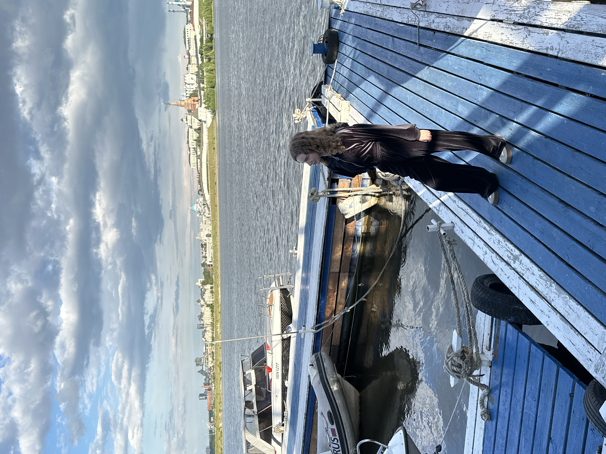 A woman stands on a blue wooden pier near docked boats on a body of water. The cityscape of Kazan, Russia is visible in the background. The sky is cloudy.