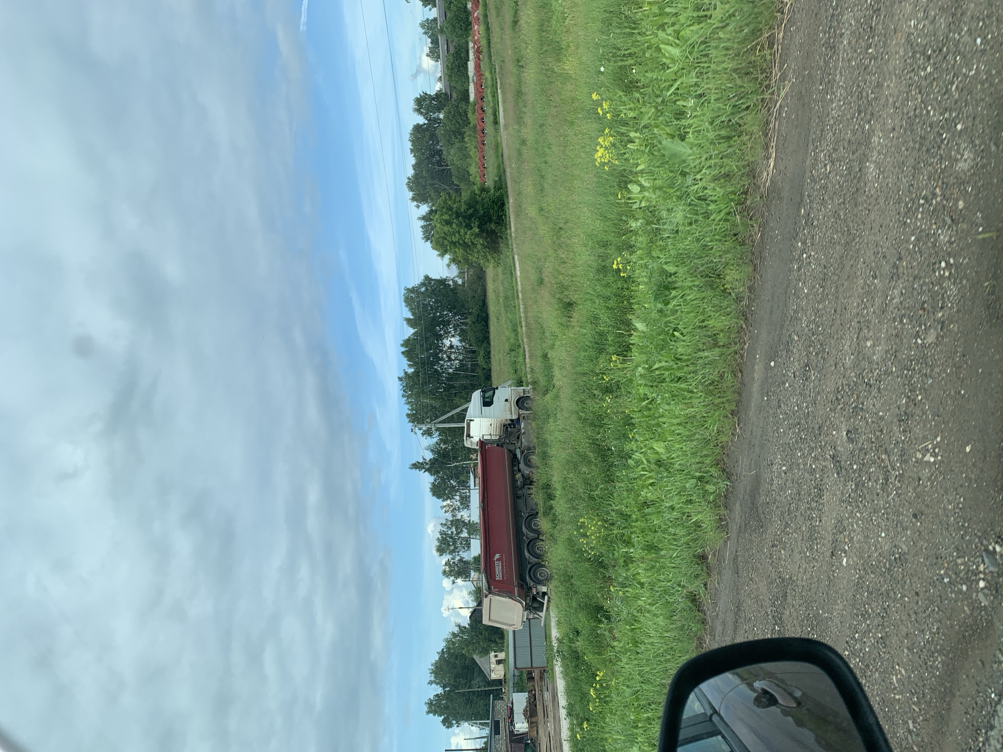 The image depicts a rural scene, likely in the city of Orda, Russia, under a cloudy sky. A large, red dump truck with a white cab is parked on a dirt road, partially covered by tall green grass and wildflowers. The truck has multiple axles and appears to be a commercial vehicle. In the background, there are several buildings, including what looks like a workshop or storage area with metal structures and a small, older building. A line of trees borders the field, and in the distance, power lines are visible. The perspective suggests the photo was taken from a moving vehicle, as evidenced by the side mirror in the lower left corner of the frame. The overall impression is one of an industrial or agricultural area on the outskirts of a settlement.