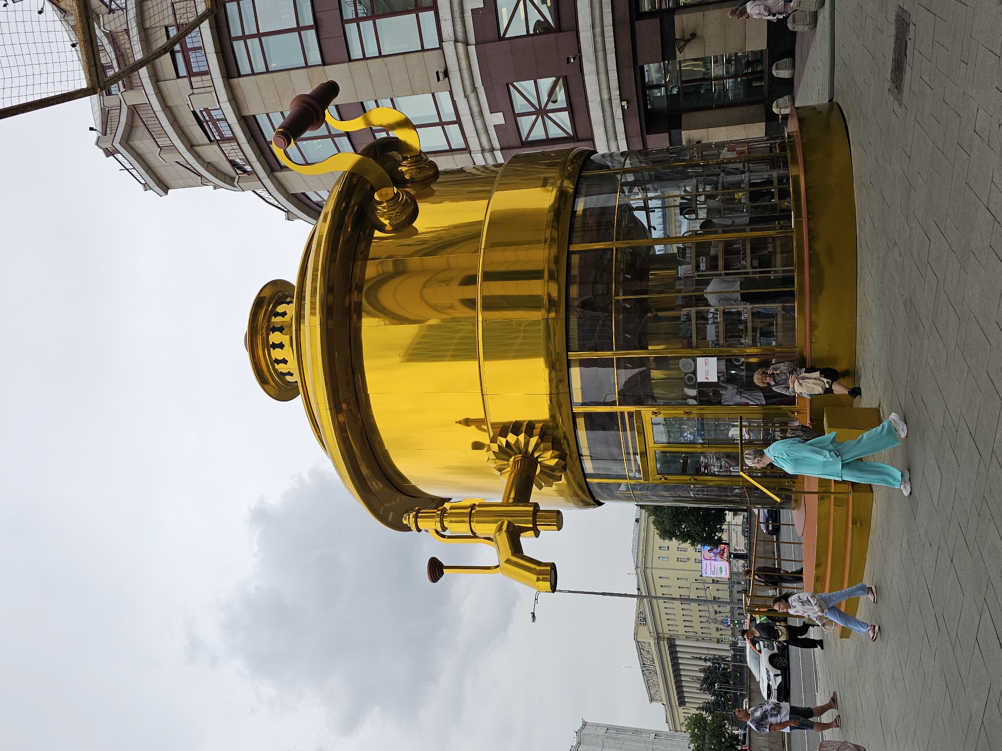The image captures a vibrant outdoor scene in Moscow, Russia, dominated by a large, golden, cylindrical structure that resembles a faucet or a vintage industrial machine. This striking object has a glass enclosure, revealing shelves and items within, possibly a shop or exhibition. A stylized, golden faucet-like appendage extends from its side. The structure is positioned on a paved pedestrian area with several people walking around it and interacting with the environment.
In the background, there are multi-story buildings with modern and classical architectural elements, characteristic of urban Moscow. The sky is overcast, suggesting a cloudy day.
Several individuals are visible in the frame. One person in a bright turquoise outfit is walking alongside the golden structure. Further along the sidewalk, a group of people are walking, including a person in a white top and blue jeans, and another in dark clothing. Towards the bottom right, a person in a dark shirt and shorts is standing. Other figures are seen within or near the golden structure.
The overall atmosphere appears to be that of a public space, possibly a square or a busy street, where people are going about their day. The prominent golden object draws attention, suggesting it might be a landmark, an art installation, or a unique commercial space. The time of day appears to be daytime, given the lighting conditions.