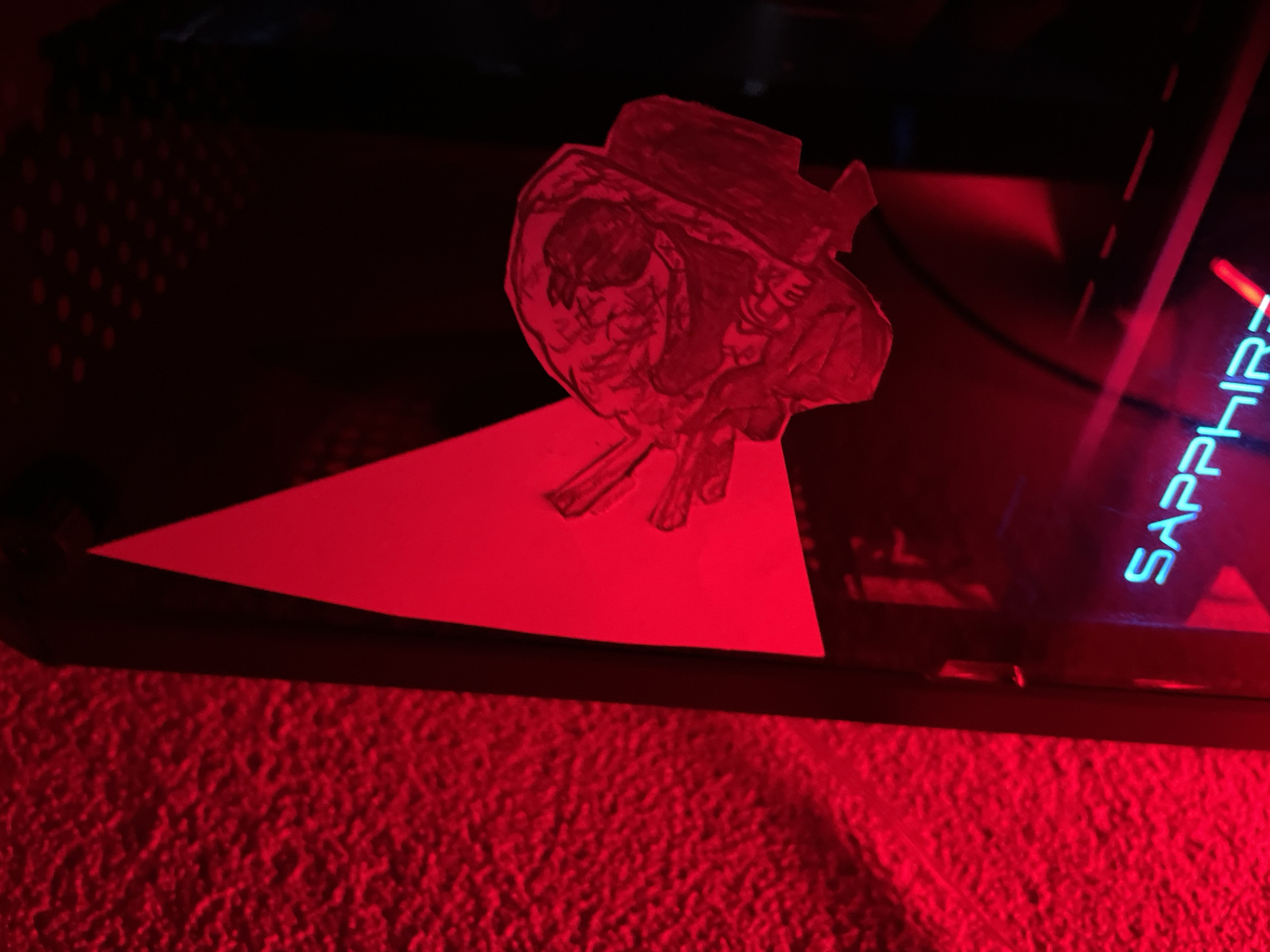 The image depicts a hand-drawn, crudely cut-out paper representation of a human heart wearing a hat, seemingly placed on top of a triangular piece of red paper. This is positioned on a dark, possibly metallic, surface. To the right of the heart is a neon-lit word "SAPPHIRE". The background and foreground are largely illuminated by a strong red light, giving the entire scene a monochromatic red hue. No specific location in Zlatograd, Bulgaria can be determined from this image.