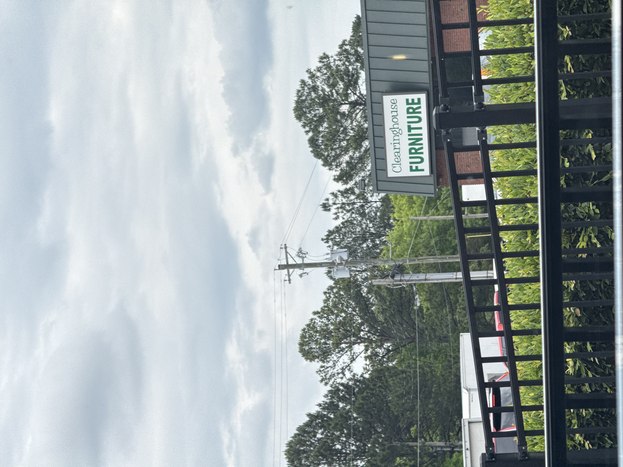 The image depicts a roadside scene near Stone Mountain, Georgia. The focal point is a sign for "Clearinghouse Furniture," mounted on a building behind a dark metal fence. Power lines and a utility pole rise in the background, partially obscured by trees. A glimpse of a white truck or trailer with red accents is visible behind the fence and foliage. The sky is overcast with heavy clouds.