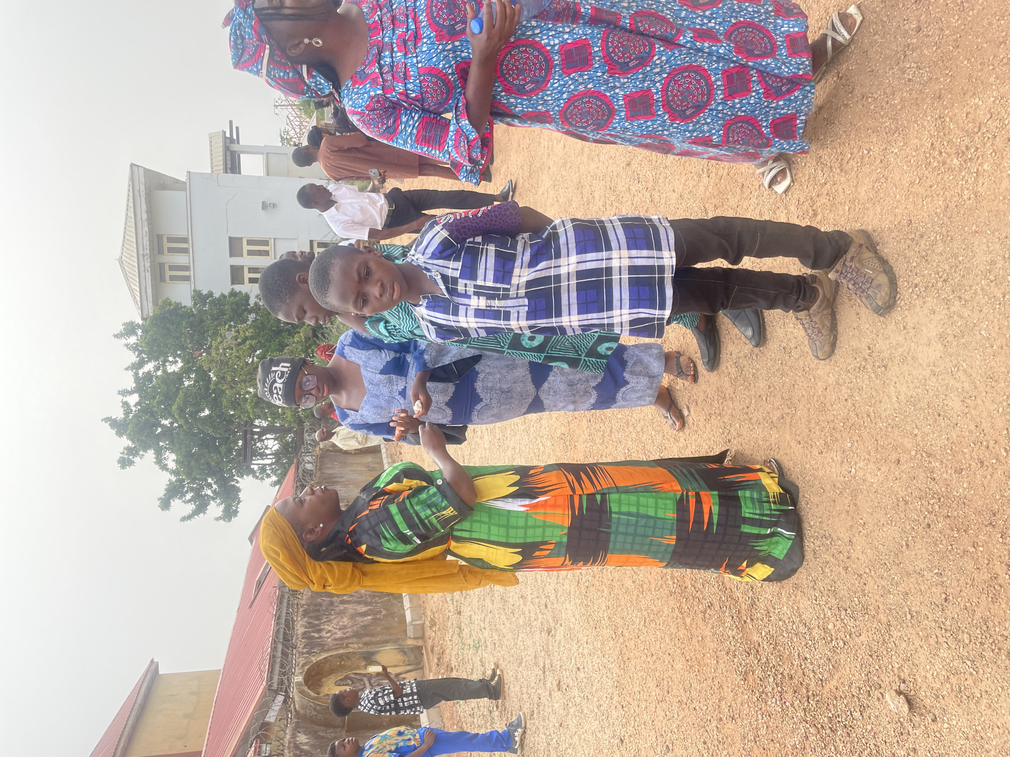A group of people stand outside on a ground of compacted dirt with scattered pebbles. The location is Jos, Nigeria. A child stands in front, wearing a blue and white plaid shirt and black pants. The woman at the bottom of the frame wears a yellow hijab and a long dress with a bright, colorful pattern. The other people in the image have various attire. A building can be seen behind the group, along with some trees. The sky is overcast.