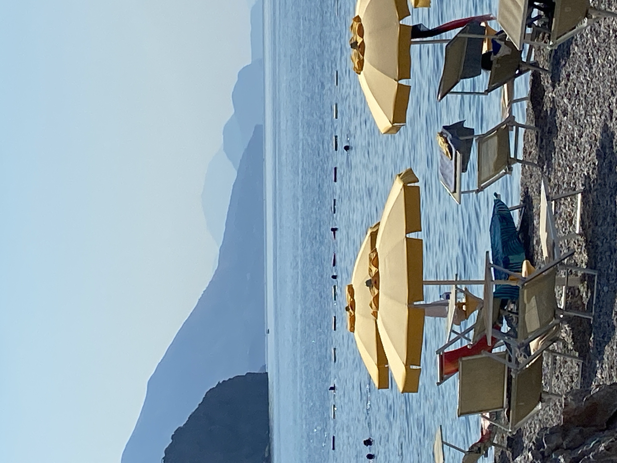 The image depicts a serene beach scene in Prijevor, Montenegro, on a clear, sunny day. The foreground is occupied by a gravelly beach with several empty beach chairs and yellow umbrellas, some of which are open. The sea, a deep blue, stretches towards the horizon, where layers of mountains are visible, fading into a hazy blue in the distance. A rope barrier with buoys marks a section of the water, and several people are seen swimming or wading within this area. The overall atmosphere is peaceful and inviting, suggesting a typical summer day at a coastal resort.