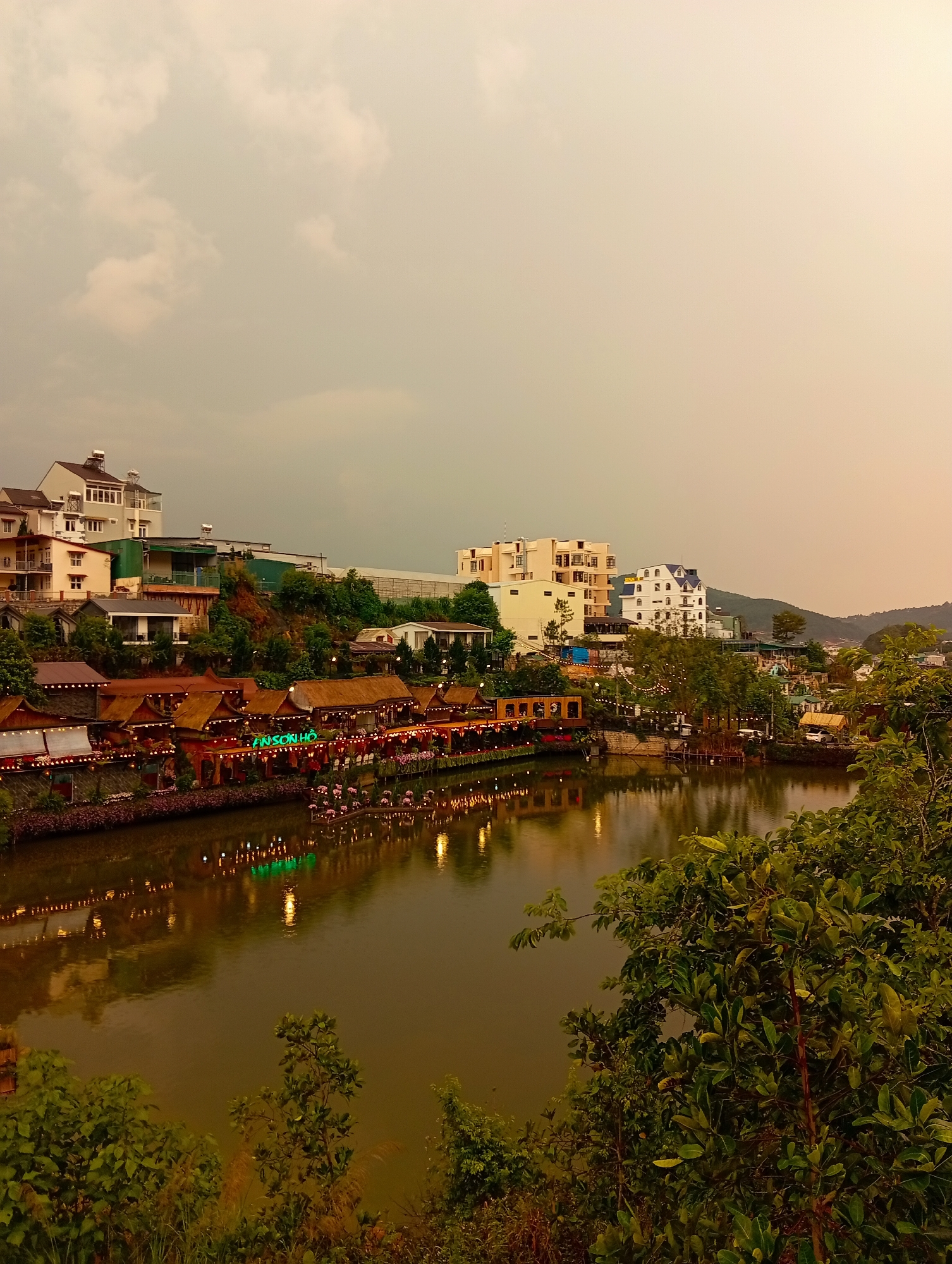 The image captures a scenic view of a lake surrounded by buildings in Da Lat, Vietnam. The sky is overcast, casting a diffused light over the scene. A row of restaurants with thatched roofs lines the edge of the lake, adorned with lights that reflect in the water. The reflection creates a symmetrical visual effect. The structures are constructed on the banks of the lake.
The lake water is calm and reflects the surrounding structures. Buildings of varying heights and architectural styles stand on the hillside behind the restaurants. Trees and greenery are visible on the banks of the lake, partially obscuring the view in the foreground.