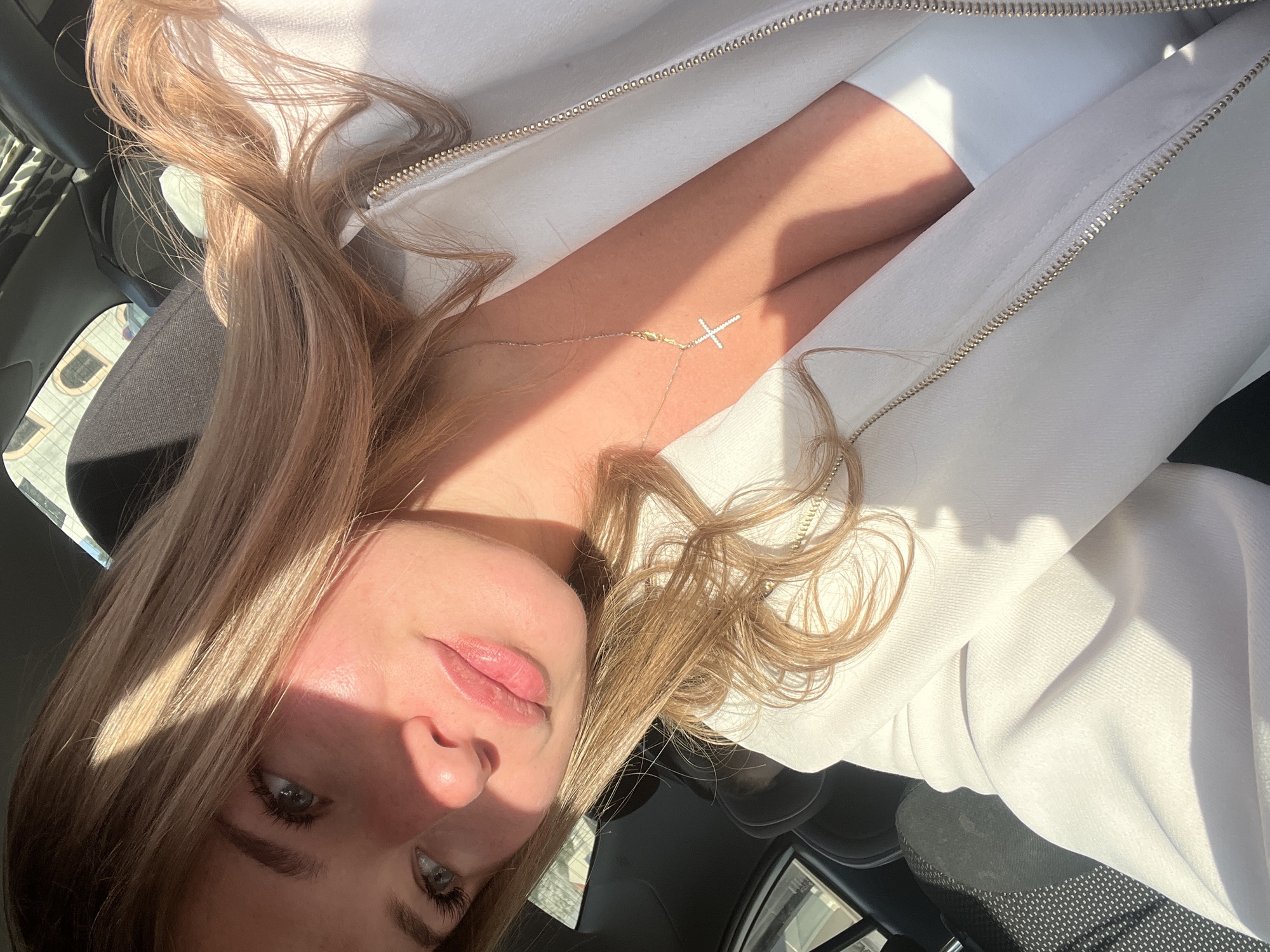 A portrait of a young woman with light-brown wavy hair is taken inside a car. She is wearing a white jacket and a delicate necklace with a cross pendant. Her face is partially illuminated by sunlight. The interior of the car is visible, including the dashboard and seat. The location is Budva, Montenegro, as indicated by the window view. The environment shows a city setting.