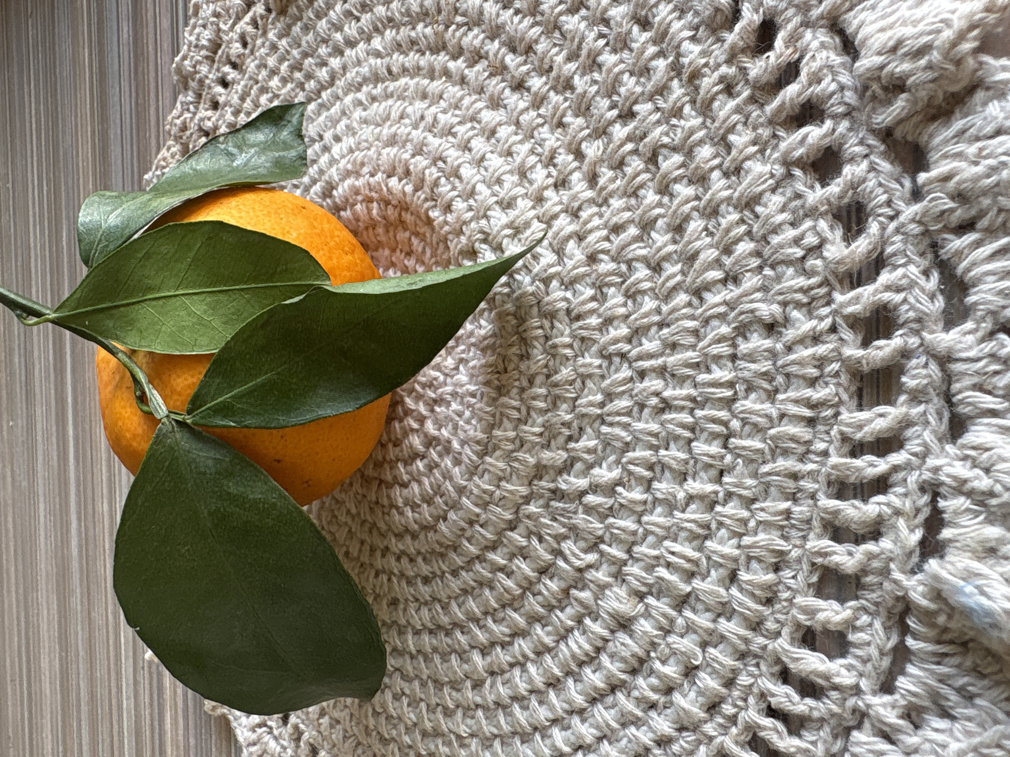 A close-up, overhead shot shows an orange with green leaves resting on a crocheted placemat. The placemat is beige and has a circular, concentric pattern with a lacy edge. The background appears to be a wooden surface. The image is a still life. The location is not identifiable within the city of Bratsk, Russia.