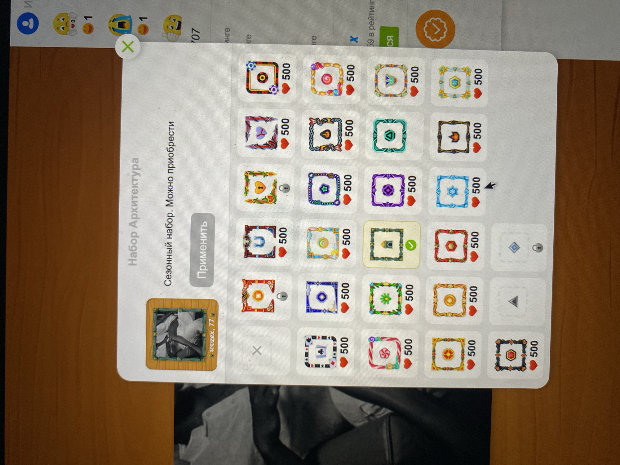 The image shows a screenshot of a user interface, likely from a game or application. The UI displays a selection of decorative frames or borders, each with a heart icon and the number "500" below it, indicating a cost or requirement. The title at the top reads "Набор Архитектура" (Set Architecture). Below this, there is text that translates to "Seasonal set. Can be purchased." A button labeled "Применить" (Apply) is visible.
The frames are displayed in a grid, with various designs including geometric patterns, symbols like a lock, a star, a gem, and a crown. One frame is currently highlighted with a green checkmark, suggesting it is selected or already in use.
The overall scene is a digital interface on a screen, with the user's desk surface visible at the bottom. The context of Khankurgan, Uzbekistan, is not depicted in the image.