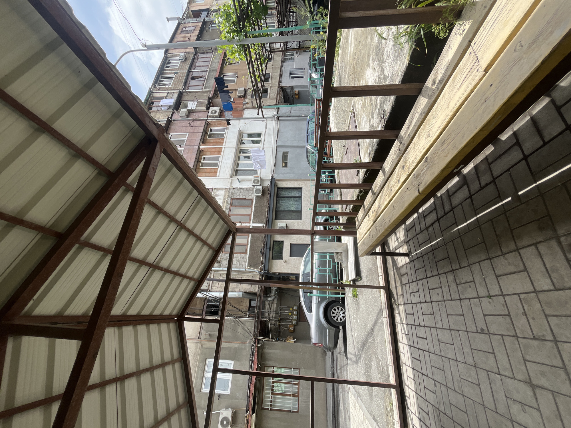 The image depicts a scene in Yerevan, Armenia, viewed from under a metal-framed structure with a corrugated metal roof. Adjacent to the structure is a wooden bench overlooking a paved area. Beyond the paved area, a silver car is parked in a narrow street lined with aging, multi-story apartment buildings. Clotheslines with hanging laundry are visible between the buildings. Air conditioning units are affixed to the building facades. The sky is partially visible above the buildings. The image appears to be taken during daylight hours.