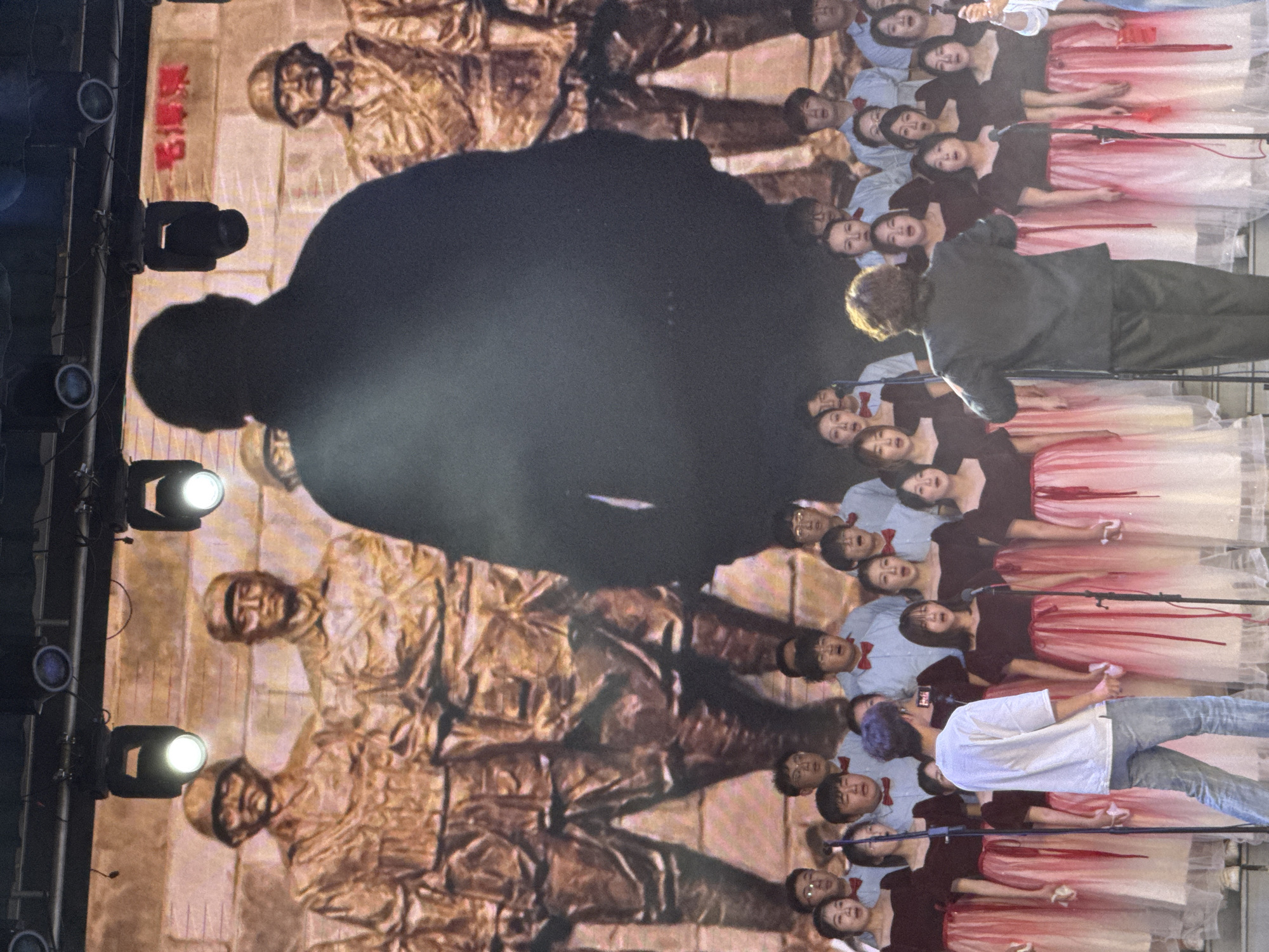 The image depicts a vibrant indoor performance, likely a choral concert or cultural event, set in Guta, China. The scene is illuminated by artificial lighting, with bright stage lights visible on metal trusses along the left side.
In the foreground and mid-ground, a large choir of young individuals is arranged in rows, with many faces directed upwards and mouths open, suggesting they are singing. Some choir members wear light blue shirts with red bow ties, while others are dressed in dark tops paired with long, flowing skirts that feature a white or light-colored base fading into red or pink. Microphones on stands are interspersed among the performers.
A person with curly hair, possibly a conductor, is visible from behind in a dark suit, facing the choir. To their right, another individual with purple hair, wearing a white t-shirt and blue jeans, is seen from behind, holding what appears to be a mobile phone or small camera, seemingly recording the event. A large, dark silhouette of a person, likely an audience member or stage crew, partially obscures the upper left part of the background.
The background is dominated by a massive screen displaying a historical or patriotic image. This image resembles a bronze-toned mural or depiction, featuring multiple figures in what appear to be military uniforms, possibly representing soldiers or historical heroes. Visible on this screen, near the top left, are red Chinese characters, which are difficult to fully discern but include "革命" (gémìng), suggesting "Revolution" or related historical text. The overall atmosphere appears focused and engaging, indicative of a formal performance.