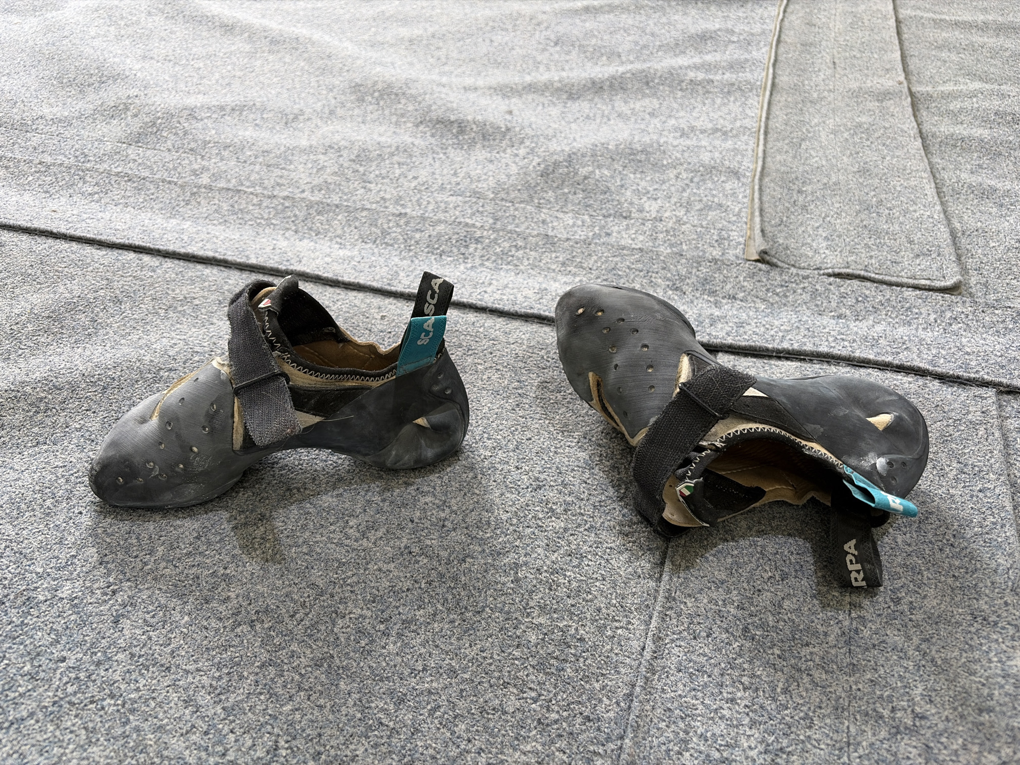 The image shows a pair of used climbing shoes on a gray, textured surface, likely a climbing gym mat. One shoe is oriented with the heel facing the viewer, displaying the brand "SCARPA" on the pull tab. The other shoe is positioned with the toe facing towards the top right corner of the image and has "RPA" printed on its pull tab. The scene is devoid of people or other objects. Given the textual information provided, the location is assumed to be in a climbing facility somewhere in Ipswich, United Kingdom.