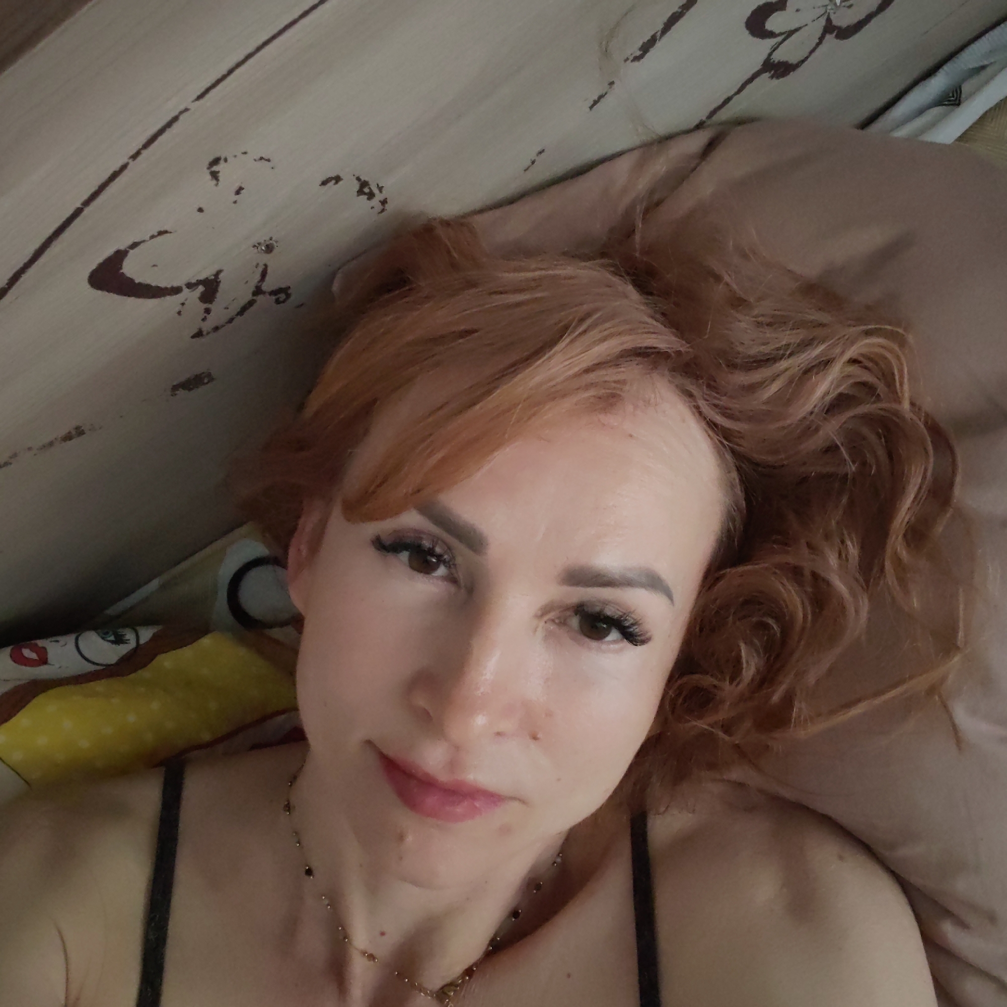 A woman with reddish-blonde curly hair is lying on a bed. She has fair skin, brown eyes with false eyelashes, and pink lips. She is wearing a black top with thin straps and a gold necklace. Her head is resting on a beige pillow. Next to her is a bed with cartoon figures in front of a wooden headboard with dark designs. The location is likely indoors. The city is not identifiable from this image.