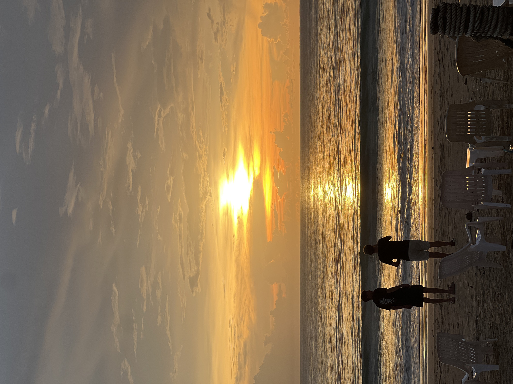 The image shows a beach scene at sunset. The sky is filled with golden clouds, and the sun is low on the horizon, reflecting off the water. Two people are standing on the beach near the water's edge. Several plastic chairs are scattered on the sand nearby. The location is Unknown, Unknown.