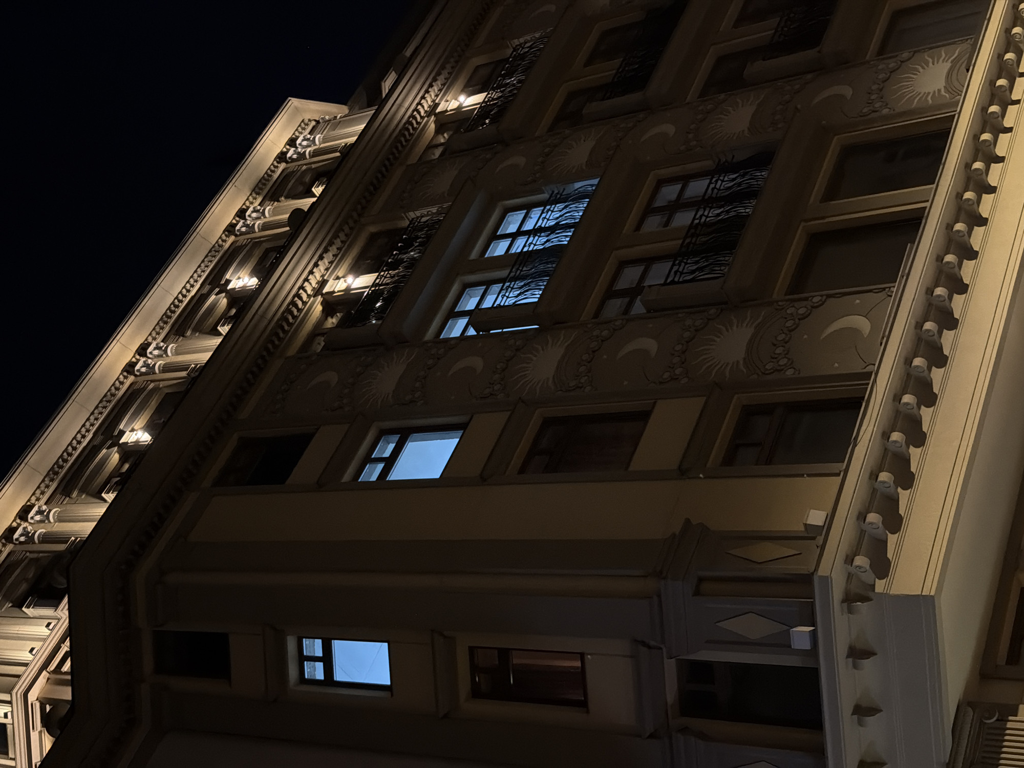 The image shows the façade of a building at night. The building has an ornate exterior, with decorative details like stylized sunbursts and geometric patterns. Many windows are visible, some with light emanating from within, indicating the presence of interior lighting. The lighting on the building's exterior highlights its architectural features. The sky is dark, suggesting it is nighttime. The location appears to be urban, but without street signs or other contextual clues, it is impossible to confirm the Moscow, Russia location.