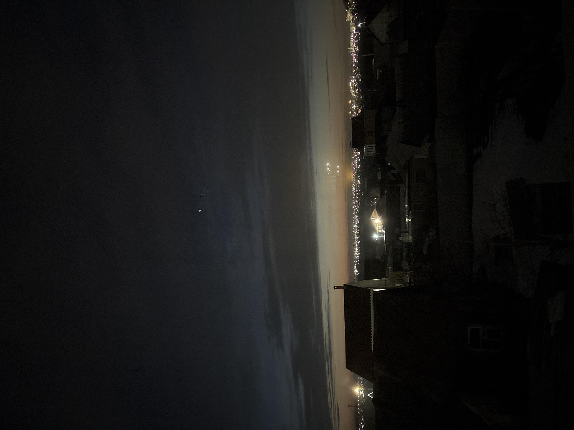 The image depicts a nighttime cityscape, potentially of Ulyanovsk, Russia. The view is from an elevated position, likely a building or hill. To the right, dark buildings and trees are visible, with a structure featuring a lit window in the foreground. A faint line of lights extends across the horizon, indicating urban development along the Volga River. The left side of the image shows a dark, cloudy sky.