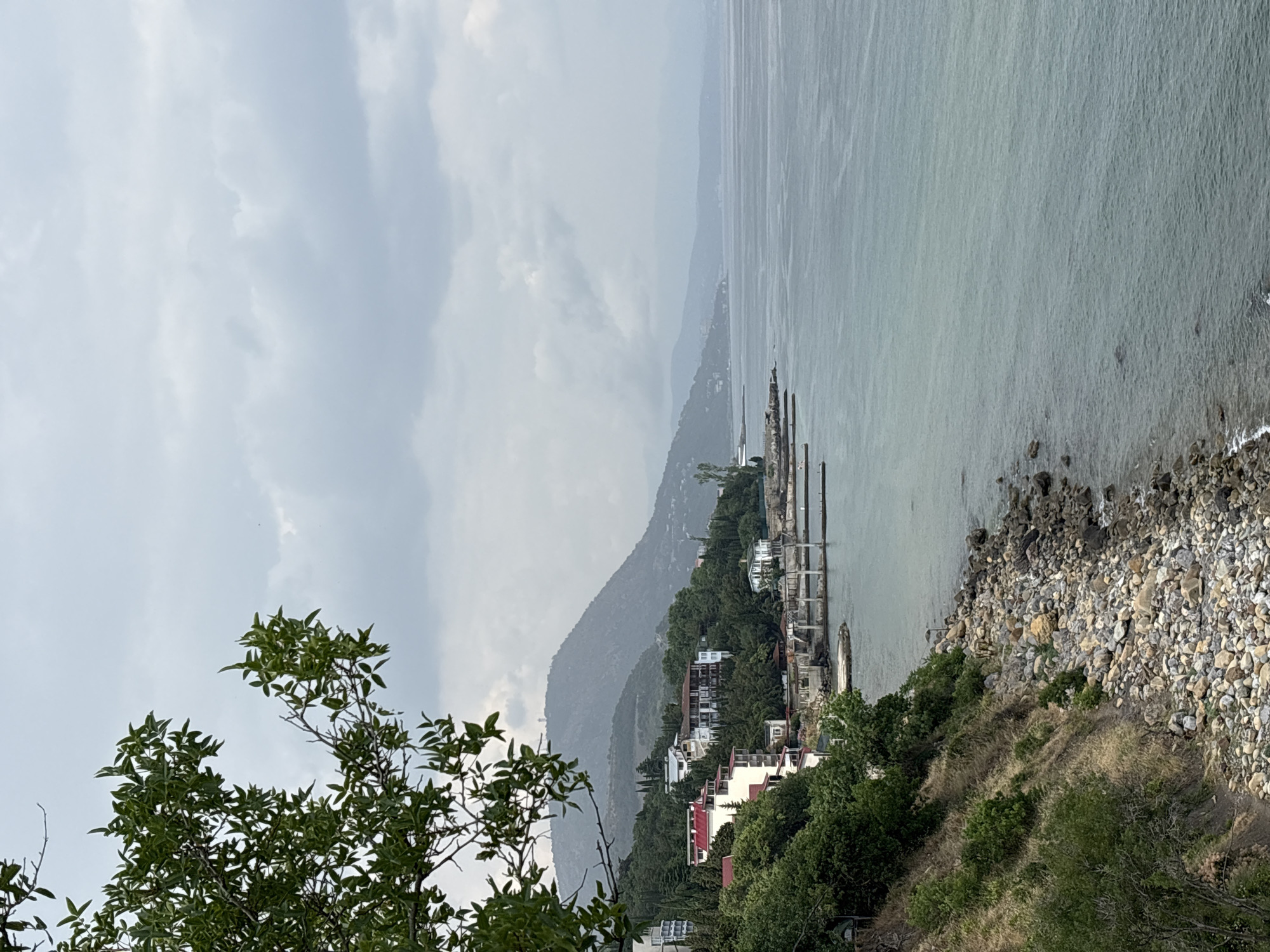 This is a medium shot, taken from an elevated position, of a coastal scene in Bondarenkovo, Ukraine. In the foreground, on the left, a tree branch with green leaves hangs down from the top of the frame. Below the tree, a steep, rocky embankment covered in dry grass and scattered greenery slopes down towards the sea. The shoreline is composed of numerous rounded pebbles and rocks.
Further into the scene, along the coast, a series of buildings with red and white roofs are nestled amongst lush green trees. Some of these appear to be houses or small hotels. There are also several piers or jetties extending into the water.
The sea occupies the right and central portion of the image, appearing calm with a muted, greenish-blue hue under an overcast sky. In the distance, rolling hills covered in vegetation form the backdrop, with a hazy, mountainous landscape fading into the horizon. The sky is filled with thick, grey clouds, suggesting a muted or possibly rainy day. There are no people or animals visible in the frame. The overall impression is one of a quiet, natural coastal landscape with a small settlement.