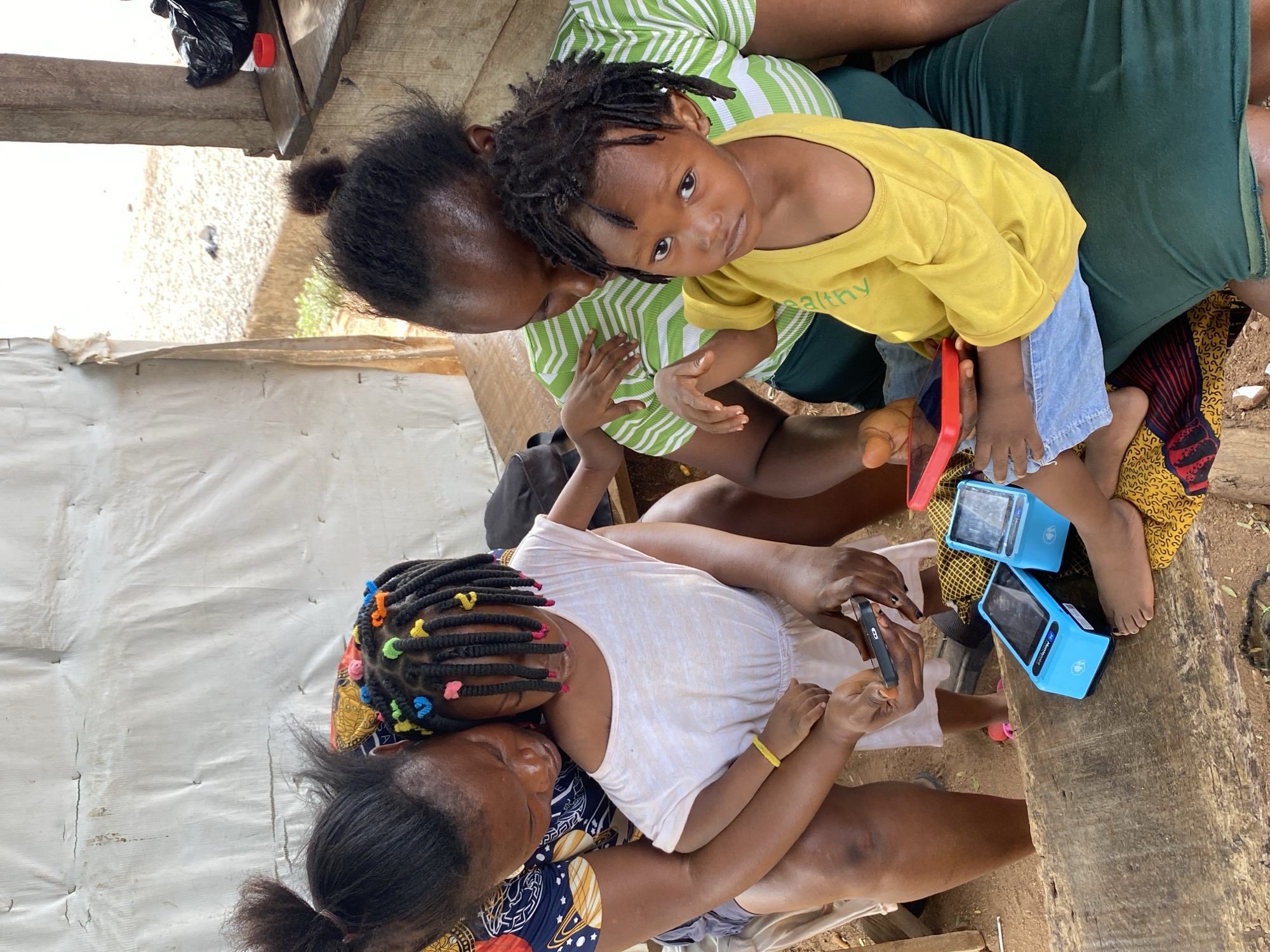Two women and two children are seated together, likely on a wooden bench. One woman wears a green and white striped shirt and dark green pants, with a young child in a yellow t-shirt with the word "healthy" printed on it, light denim shorts, and bare feet, sitting on her lap. The child looks up at the camera. The other woman wears a patterned dark blue and gold top and dark bottoms, and a younger child with colorful beads in their braided hair sits in her lap. This child wears a light-colored t-shirt and looks at a mobile phone being held by the woman. Two blue electronic devices sit near the children. The setting appears to be outdoors, in a partially covered space with wooden structures. The location is Adankolo, Nigeria.