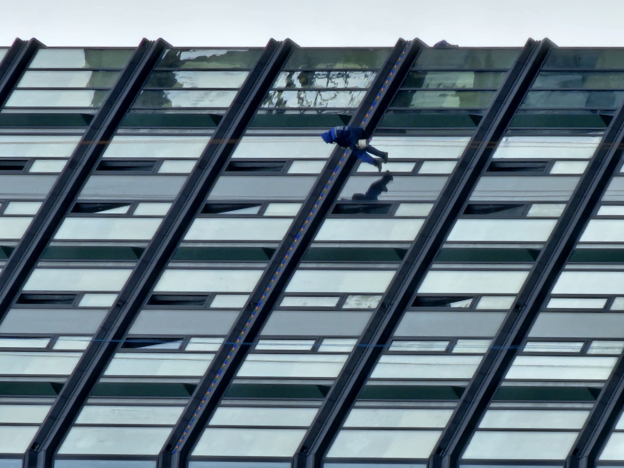 A window cleaner in a blue uniform and hood is rappelling down the side of a modern, multi-story building in Moscow, Russia. The building features a facade of dark frames and numerous large glass panels, some of which are reflecting the overcast sky. The worker is suspended by a rope and is carrying a bucket. The cleaning equipment, including the rope and harness, is visible. The overall scene depicts an urban environment with a focus on high-rise maintenance.