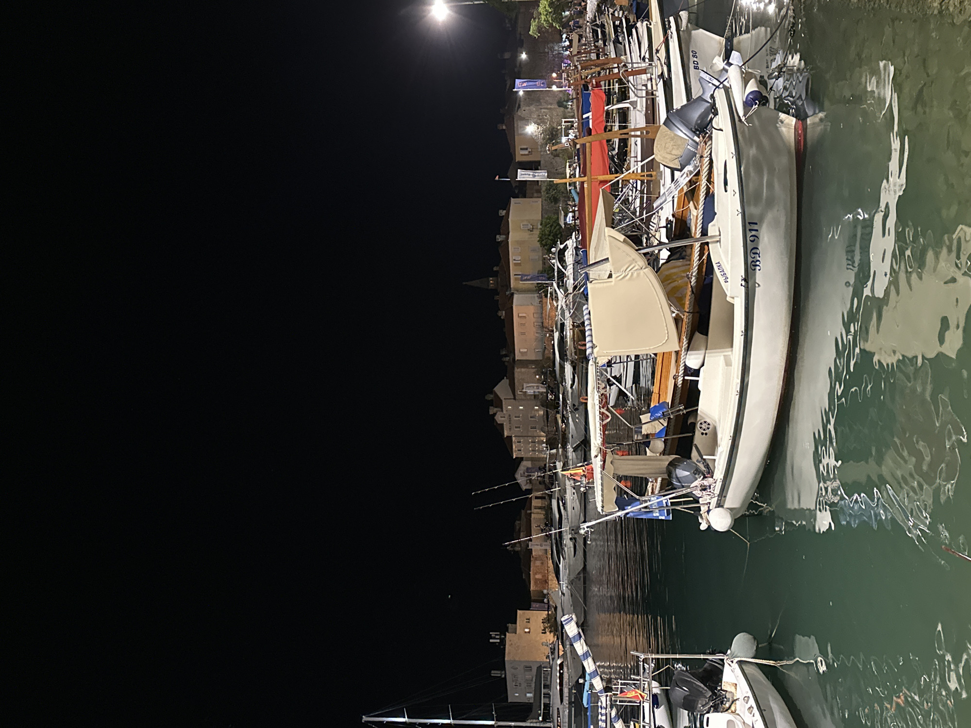 The image depicts a nighttime scene at the Budva marina in Budva, Montenegro. Several boats are docked in the water. In the foreground, a small white motorboat with a beige canopy is prominent, displaying the registration "BD 911" and "РЫБАЛКА". Another white boat is visible to its left, partially obscured. Further back, a line of larger yachts and sailboats are moored. Behind the marina, the illuminated buildings of the Old Town of Budva are visible, featuring a church spire. The sky is dark, with a single streetlamp providing some illumination on the right side of the frame. The water reflects the lights of the boats and the town. No people are clearly discernible.