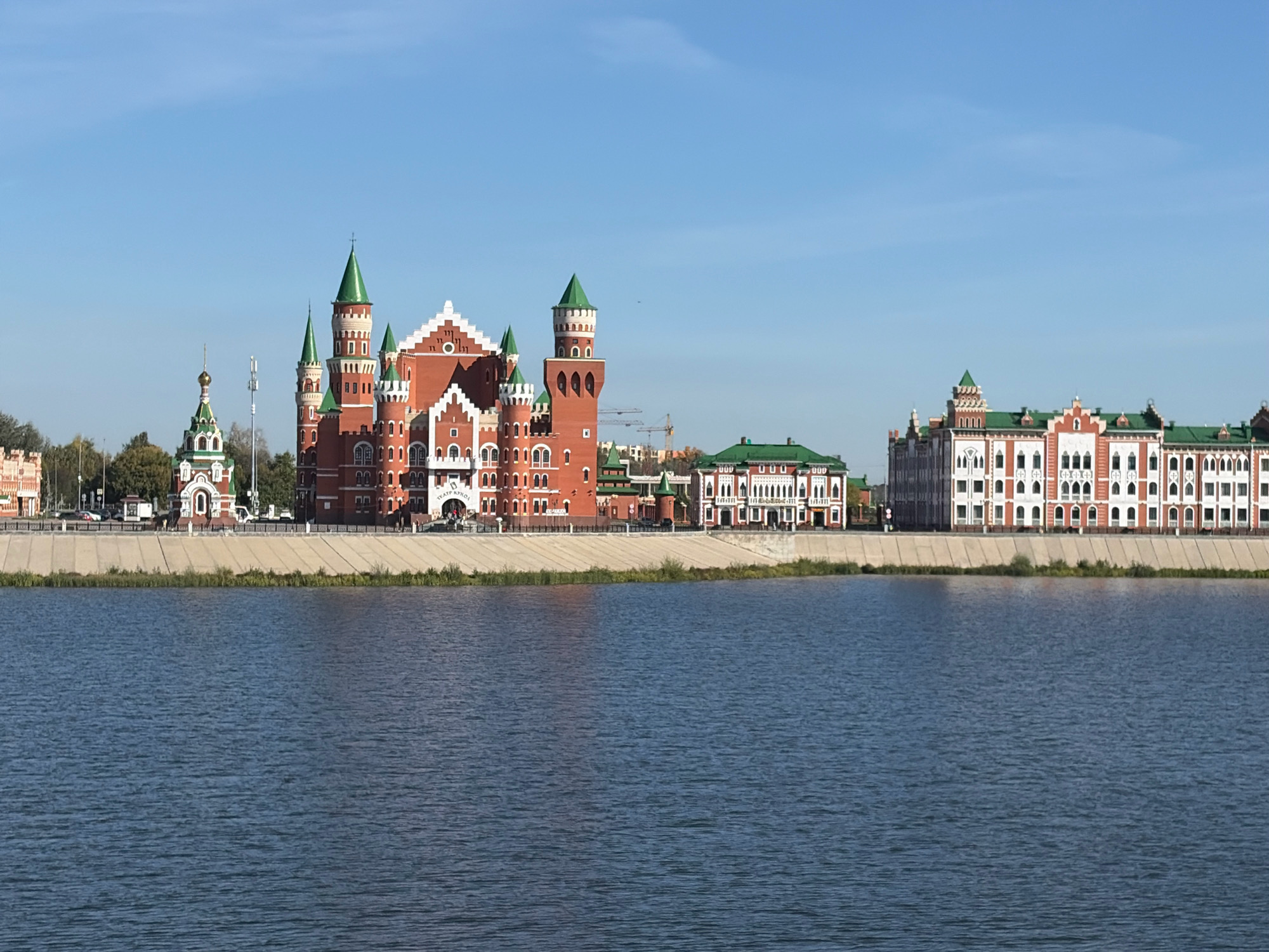 This is a wide-angle, eye-level, daytime exterior shot of the embankment in Yoshkar-Ola, Russia. The scene features a body of water in the foreground, with a stone embankment and several prominent buildings lining the opposite shore. The architecture of the buildings is varied, with some appearing to be in a neo-Gothic or castle-like style with red brick and green spires, while others are more classical with white facades and red trim. One building in the center is particularly striking with its turreted design and white-capped roofline. The sky is clear blue with a few wispy clouds. Trees are visible behind the buildings. There are no people visible in the frame. The overall impression is one of historical or stylized architecture set against a calm natural backdrop.