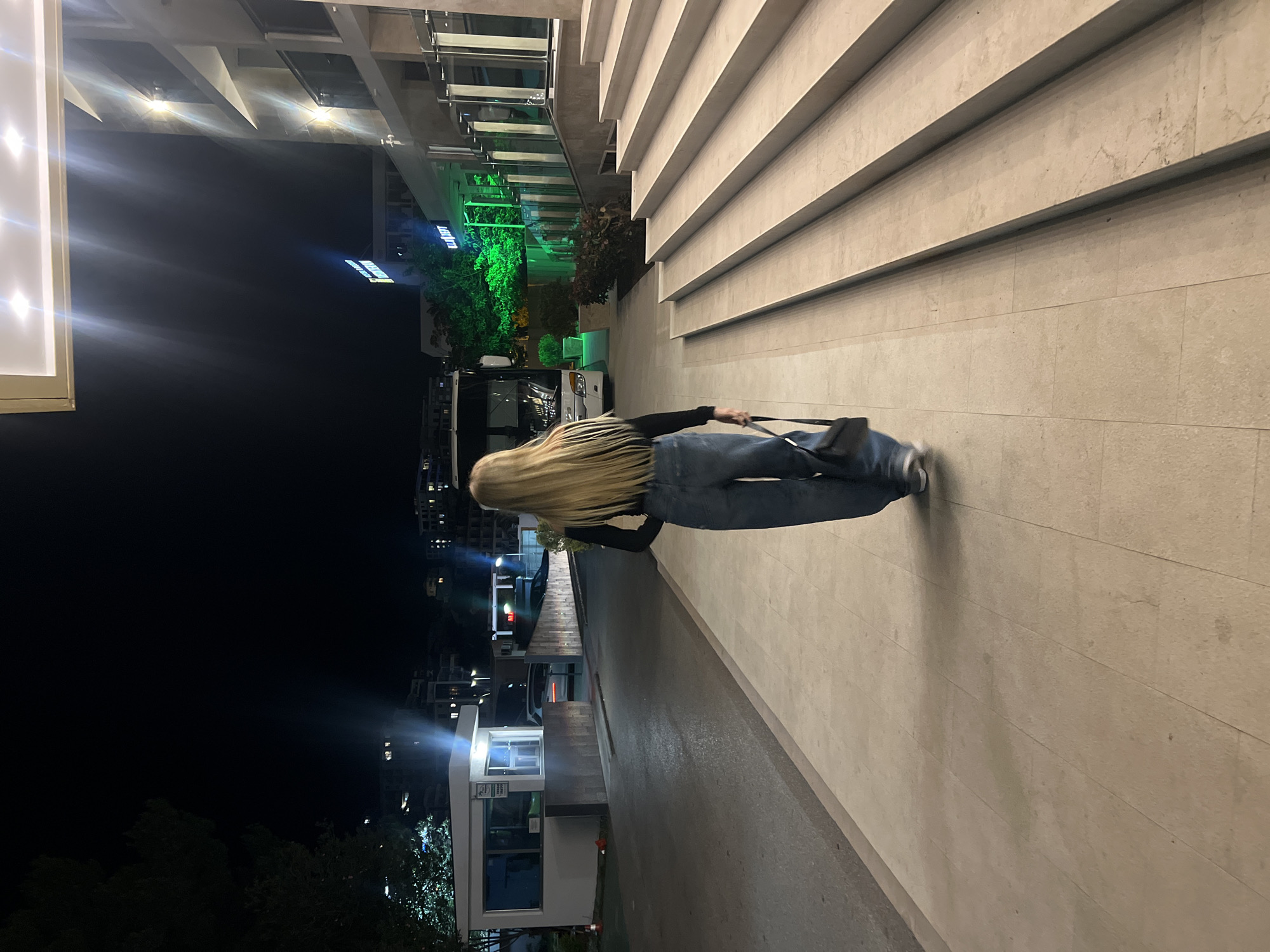 A woman with long blonde hair is walking on a sidewalk at night in Bečići, Montenegro. She is wearing dark blue jeans, a black top, and carrying a black purse. To her right is a beige building with tiered steps. To her left, across the sidewalk, is a street with various structures, including a security booth with illuminated signage. The night sky is dark, and there are other buildings and trees visible in the background.