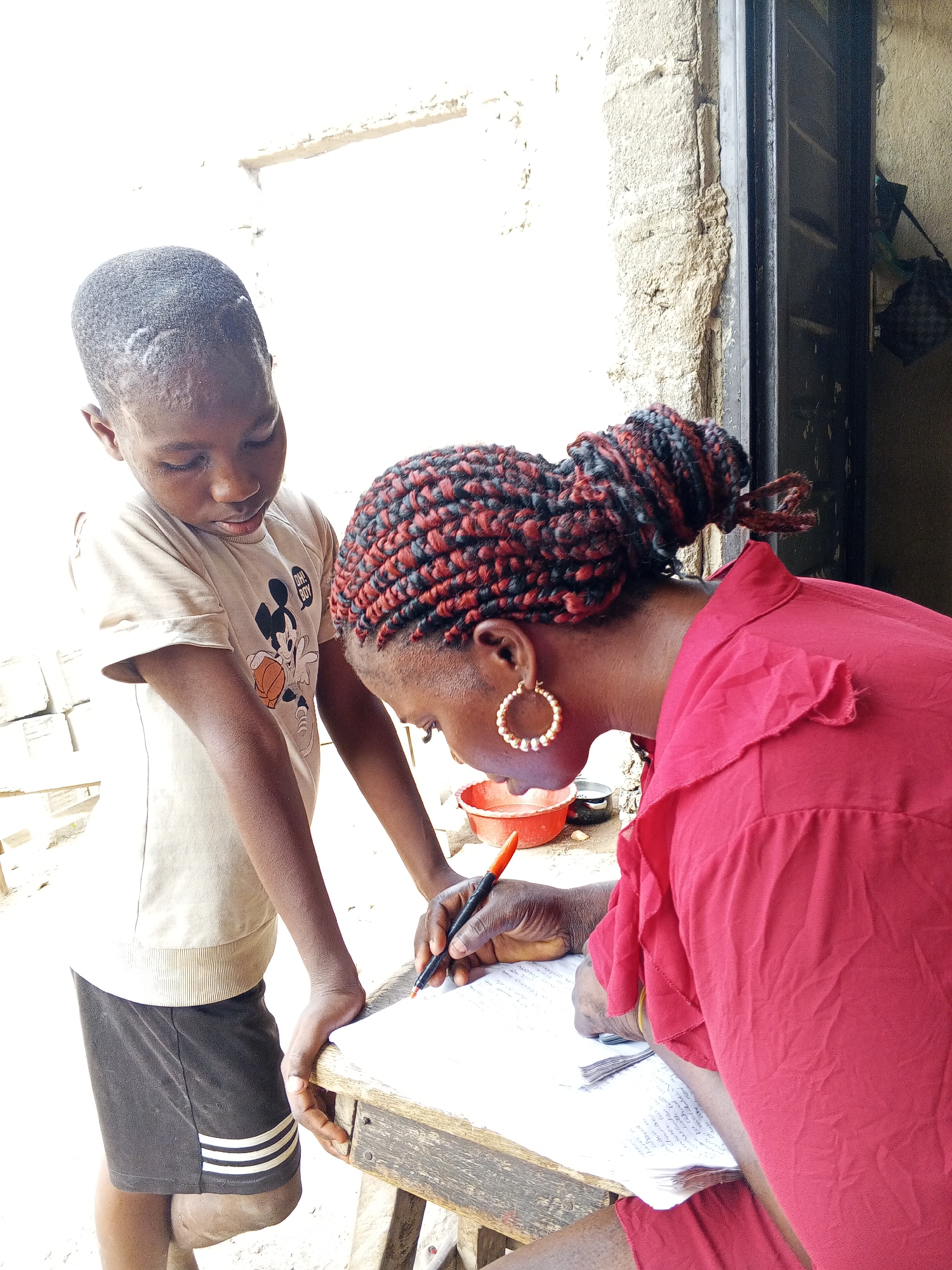 In Adonkolo, Nigeria, a young boy stands beside a woman who is writing on a sheet of paper placed on a small, weathered wooden table. The woman wears a red shirt and has her hair styled in red and black braids gathered in a bun. She is wearing gold hoop earrings and using a pen to write. The boy wears a beige shirt with a Mickey Mouse graphic and dark shorts with white stripes. They appear to be outdoors, near a wall and a doorway. A red bowl is visible on the table.
