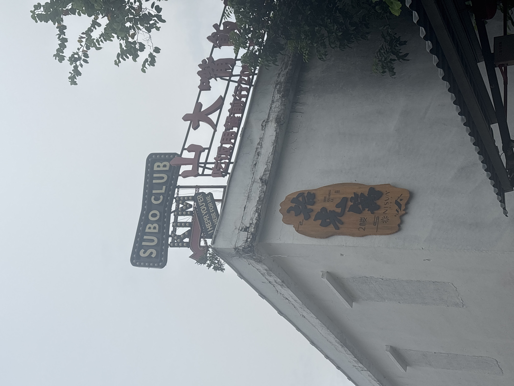 A sign on a building in Nanhai District, China, reads "SUBO CLUB" in English. Below that, another sign in English says "KIM THEPH YOU'ER HERE FOR ME". A larger sign with Chinese characters and the text "SHI GUANG LI" is also visible. The building appears to be a white, somewhat aged structure with a corrugated metal roof overhang. A tree with green leaves partially obscures the upper right portion of the image. The sky is overcast and a pale gray.