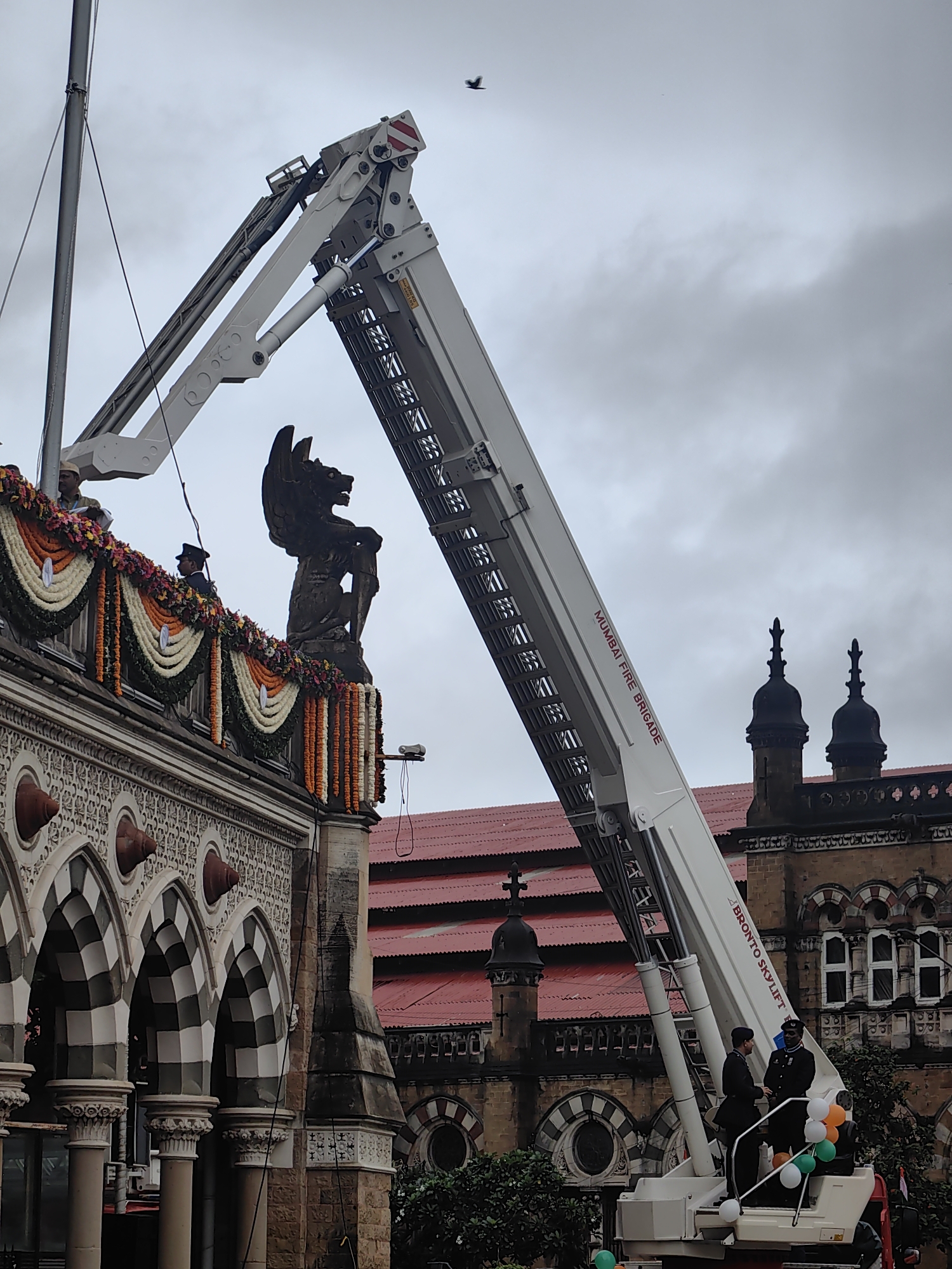 The image captures a scene outside the Chhatrapati Shivaji Maharaj Terminus (CSMT) in Mumbai, India, on a cloudy day. A large white fire truck with an extended ladder dominates the right side of the frame. The ladder is emblazoned with "MUMBAI FIRE BRIGADE" and "BRONTO SKY LIFT". Two individuals in dark uniforms, possibly firefighters or ceremonial personnel, stand in the basket at the end of the ladder, engaged in conversation. Tricolor balloons, in green, white, and orange, are attached to the basket.
The left side of the image features the iconic facade of CSMT, adorned with festive garlands of orange and white flowers. A stone sculpture of a winged creature stands atop the building's ornate structure, overlooking the scene. The building exhibits intricate architectural details, including arched windows and decorative carvings. In the background, the red-tiled roof and gothic architecture of other parts of the terminus are visible. A single bird is captured in flight against the overcast sky. The overall atmosphere suggests a formal occasion or celebration.