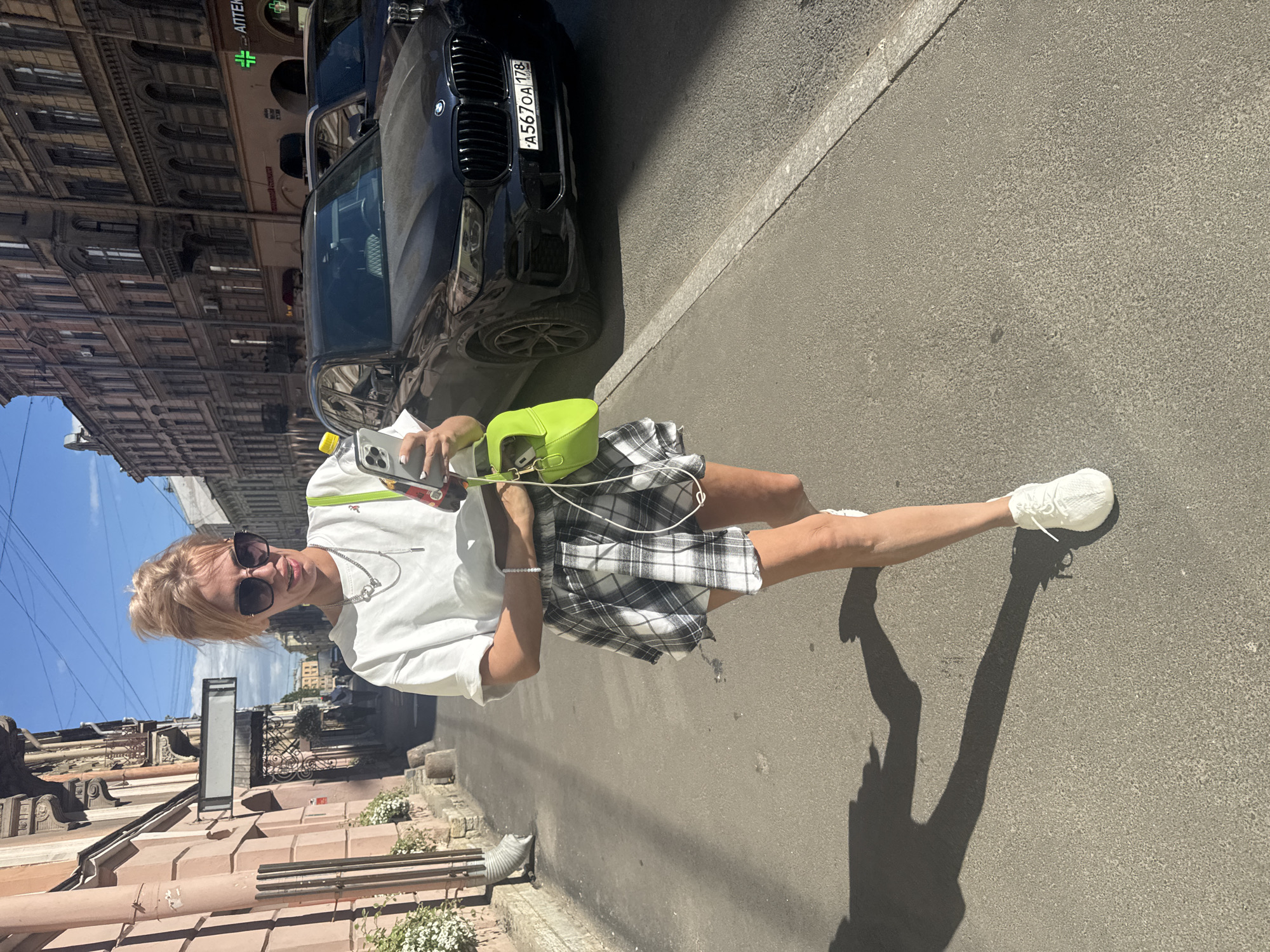 A woman with blonde hair and sunglasses stands on a sidewalk in what appears to be Saint Petersburg, Russia. She is wearing a white t-shirt, a plaid skirt, and white sneakers. She is holding a smartphone in her right hand and a water bottle with a yellow cap in her left. A bright green purse is slung over her shoulder. Behind her, a black BMW car is parked on the street. The buildings in the background are characteristic of Russian architecture, with ornate facades and tall windows. The sky is blue, suggesting a clear, sunny day. The lighting indicates it is daytime, likely midday due to the strong shadows cast by the woman and the car. The woman's expression is a slight smile, and she appears to be taking a selfie. The license plate of the BMW is partially visible and reads "A5670A 178". The scene suggests a casual outing or sightseeing in an urban environment.