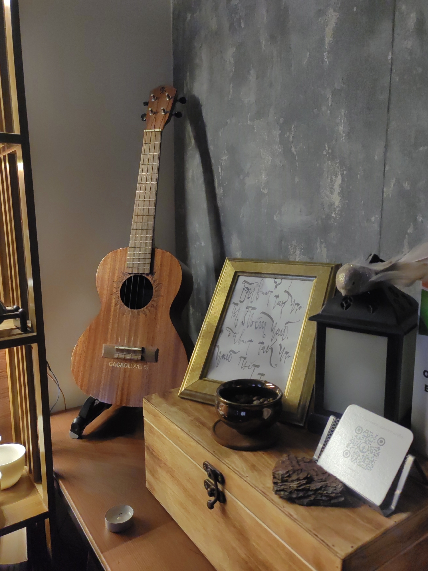 This image depicts a still life composition on a wooden surface, likely a shelf or table, set against a textured grey wall. In the background, a light-colored ukulele with "CACAOLOVER S" inscribed on its body stands propped up. To the right of the ukulele is a golden framed picture containing handwritten text in an artistic script. A dark ceramic bowl filled with what appears to be coffee beans sits in front of the frame, and next to it is a black lantern with a bird figurine perched on top. Further to the right, a white card with a QR code and text is displayed on a stand, resting on a piece of rough, dark material. A small, lit tea candle is visible on the wooden surface in the foreground. The lighting suggests an indoor setting, possibly during the day or evening, with soft light casting shadows. The overall mood is calm and curated, with a focus on decorative and musical elements. The mention of Saint Petersburg, Russia, provides geographical context but no specific visual cues within the image confirm this location.