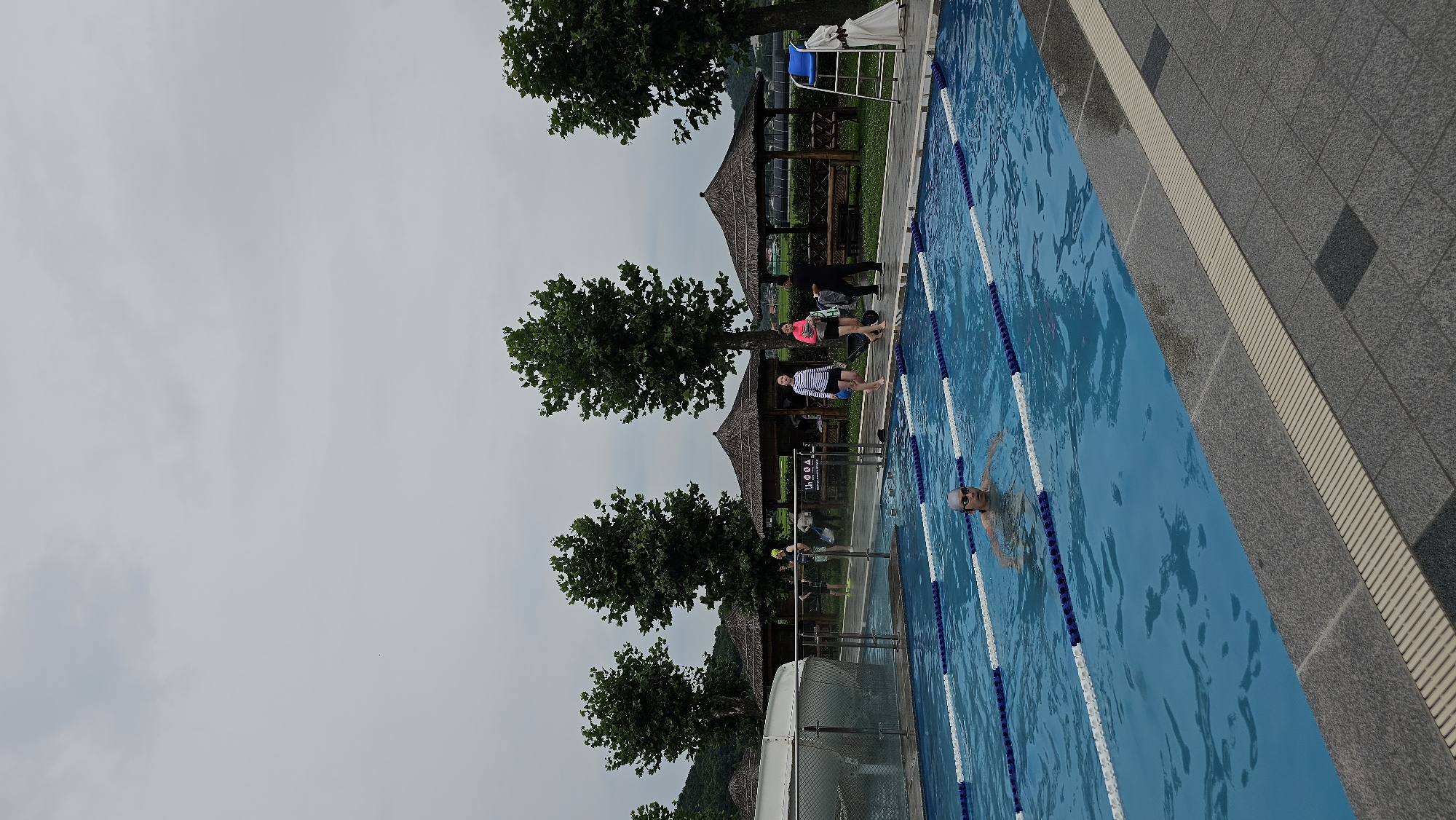A person wearing a swim cap is swimming in a clear blue outdoor swimming pool marked with white and blue lane lines. The pool is bordered by a gray stone pathway. In the background, there are thatched-roof gazebos, lush green trees, and a cloudy sky. Several people are visible on the pathway near the gazebos. A white slide is partially visible on the left side of the pool. The scene appears to be a public recreational facility in Asan-si, South Korea.