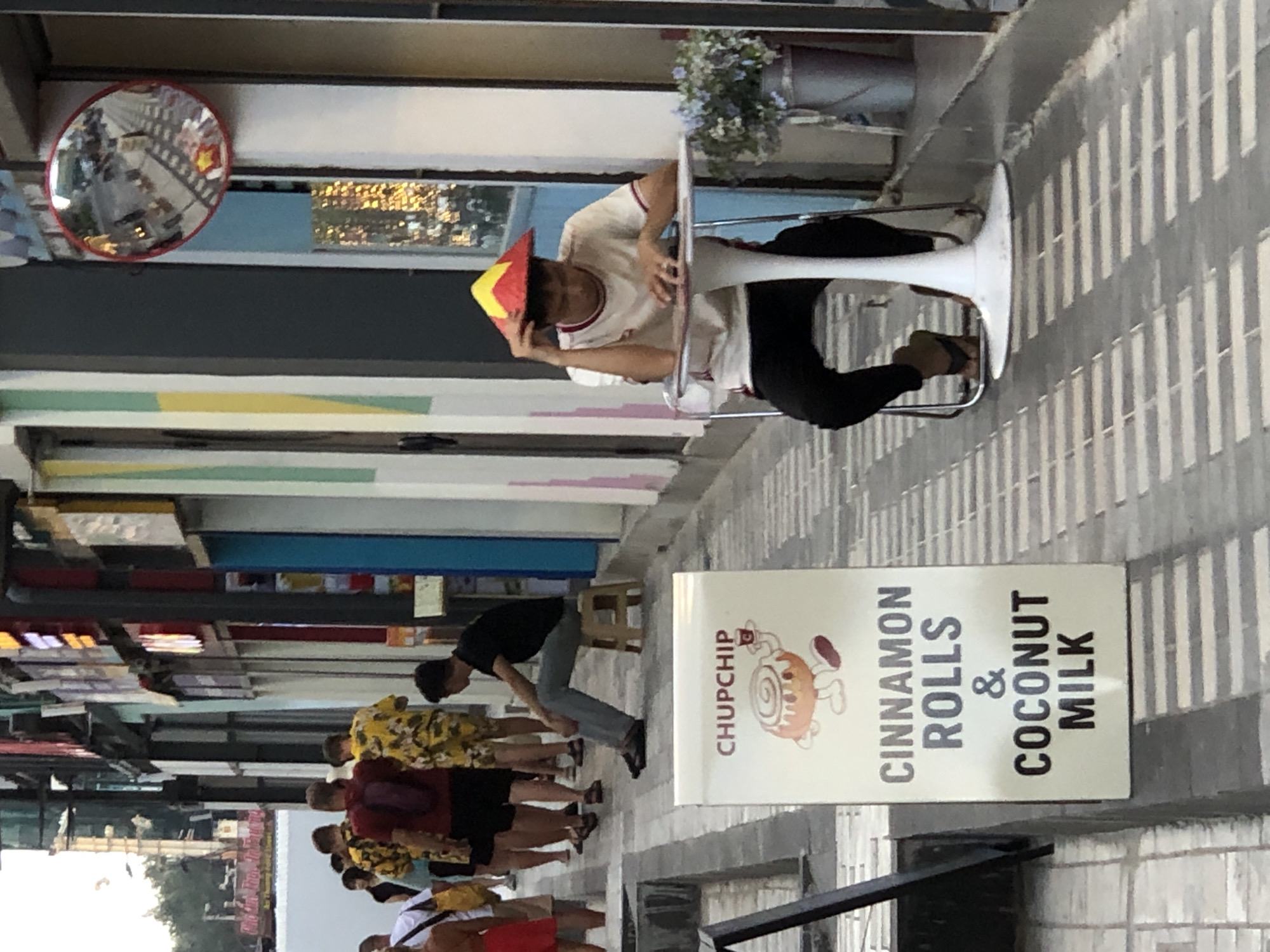 In an outdoor pedestrian walkway in Da Nang, Vietnam, a young man sits at a small white table outside a shop. He wears a white t-shirt with red trim and black pants, and a conical straw hat with red and yellow markings. His left hand rests on his forehead as he looks down, appearing pensive. To his right, a metal bucket holds an arrangement of purple flowers. In the foreground, a sign displays "CHUPCHIP" in red lettering, with an illustration of a cinnamon roll and the text "CINNAMON ROLLS & COCONUT MILK" below. A man in a black t-shirt and grey pants sits on a small stool to the left of the sign, facing away from the camera. Several other pedestrians, some wearing brightly colored tropical shirts, are visible further down the walkway. The environment suggests a commercial street with shops lining one side of the path.