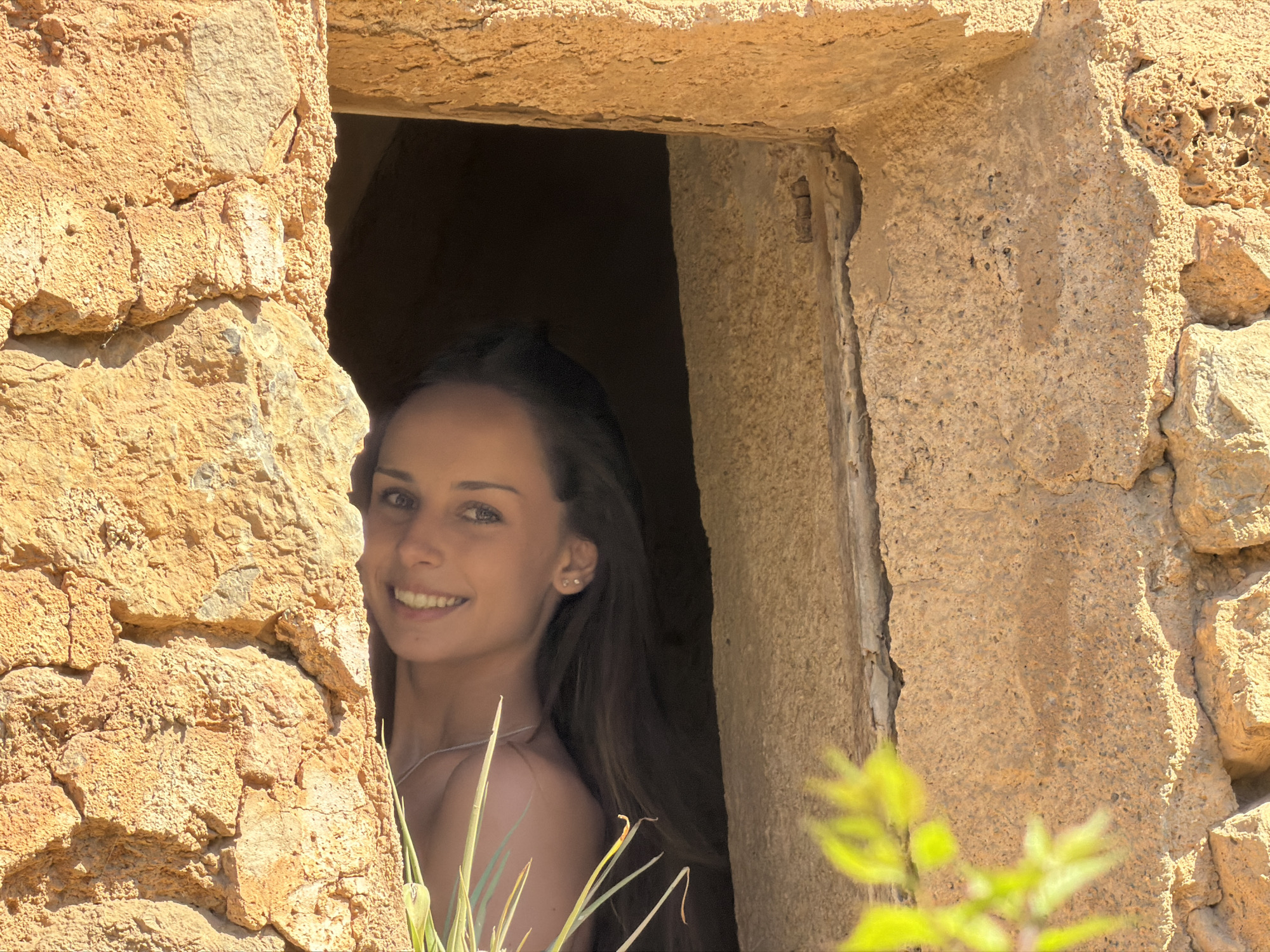 A young woman is smiling at the viewer, peeking out of what appears to be a small, rectangular stone opening. The stone structure is likely part of a building or wall, constructed from rough, light-brown rocks. The scene is sunlit, casting shadows within the opening. Green foliage is present at the bottom of the frame, partially obscuring the lower portion of the opening. No specific city information is discernible beyond the stated location in Bunyola, Spain.