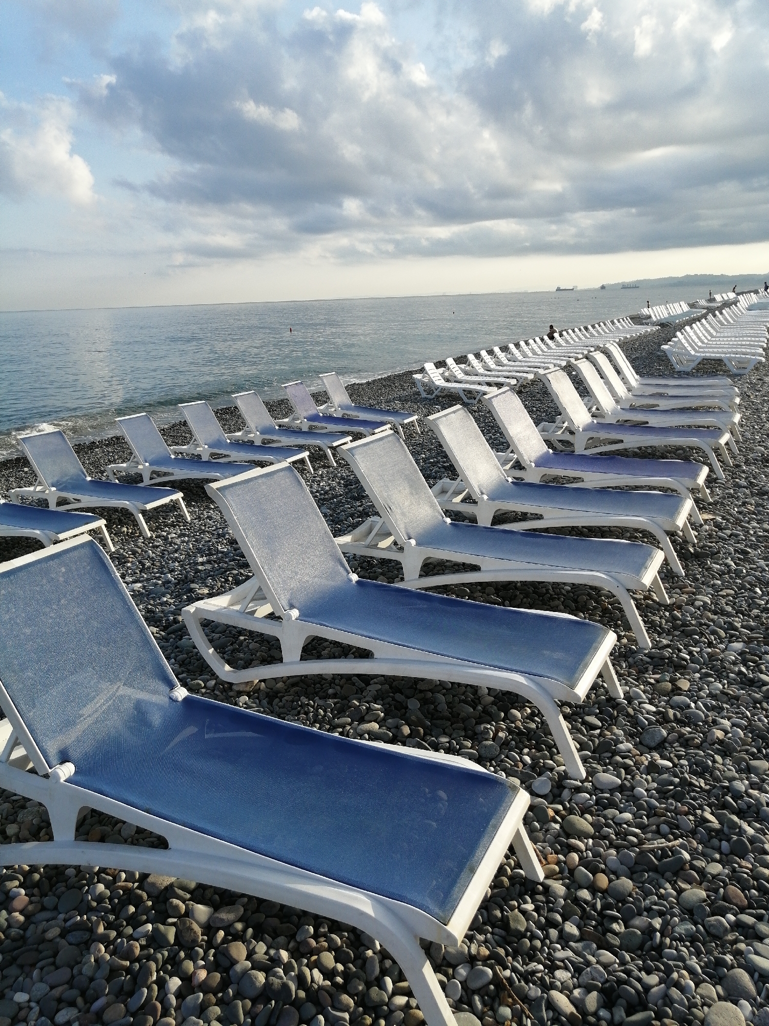 A long row of empty, blue-seated lounge chairs is arranged on a pebble beach facing a calm, grey sea. The sky is overcast with dramatic clouds. In the distance, several cargo ships are visible on the horizon, as well as a dark, hilly coastline. A few people are scattered along the beach and in the water. The scene appears to be a public beach in Batumi, Georgia, likely photographed in the late afternoon given the lighting.