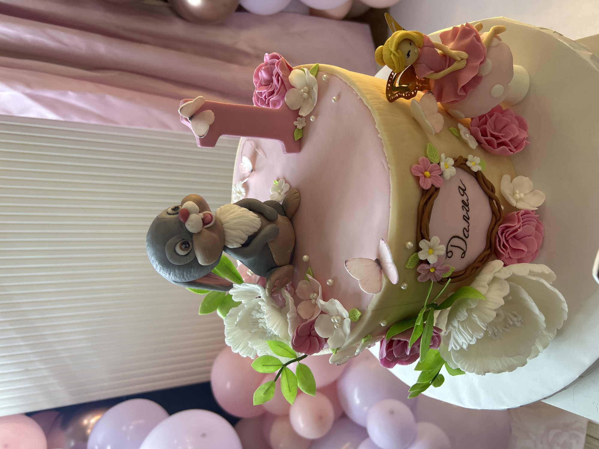 The image shows a birthday cake decorated with a rabbit figurine, the number "1," a fairy, flowers, butterflies, and a frame containing the name "Daria" written in Cyrillic script. The cake's color scheme predominantly features pink and white. The cake is placed on a table surrounded by pink and purple balloons. The location of the scene is Plovdiv, Bulgaria, based on the Cyrillic inscription.