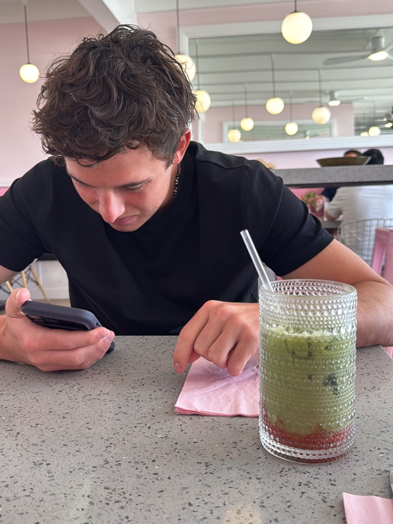 A young man in a black t-shirt is sitting at a table in a brightly lit cafe in Palm Desert, California. He is looking at his phone in his left hand while his right hand rests on a pink napkin. A tall glass of green matcha latte with a straw is on the table beside the napkin. The table is a light gray color with a speckled pattern. In the background, there are other tables and chairs, a mirror on the wall, and globe-shaped lights hanging from the ceiling.