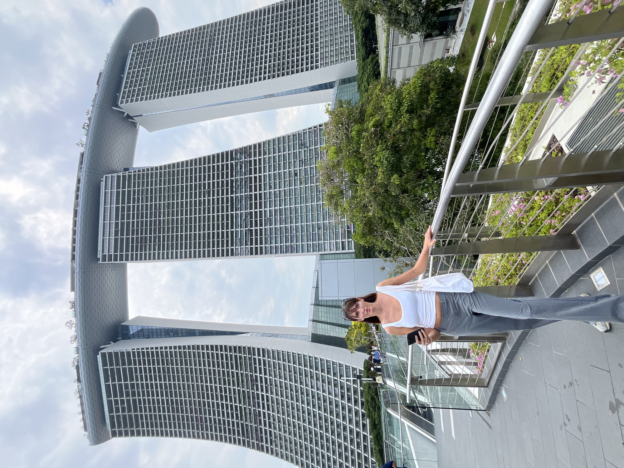 A woman stands on an elevated walkway in front of the Marina Bay Sands hotel in Singapore. She is wearing a white tank top, gray flared pants, and white sneakers. She is holding a smartphone in her right hand and has a white tote bag slung over her left shoulder. Sunglasses are perched on her head. The Marina Bay Sands hotel, a prominent landmark in Singapore, is visible behind her. The hotel's distinctive architecture features three towers topped by a boat-shaped structure. The walkway is lined with a metal railing and planters with pink flowers. The sky is overcast.