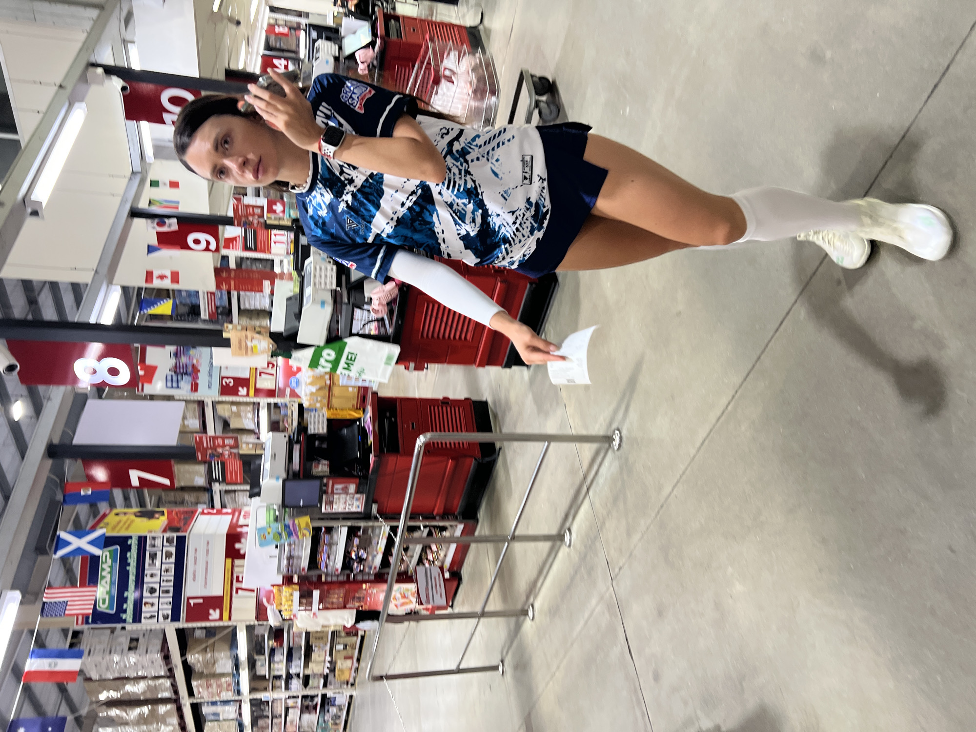 The image depicts a woman in athletic attire standing in a retail environment, possibly a warehouse store or supermarket. She is wearing a blue and white patterned athletic shirt with a logo on the sleeve, a dark blue skirt, white arm sleeves, white knee-high socks, and white athletic shoes. The woman is holding a piece of paper in her right hand and her left hand is near her ear, as if talking on the phone or adjusting an earpiece. In the background, shelves filled with merchandise, checkout counters, and promotional flags are visible. Specifically, flags of various countries and large numbered signs are hung from the ceiling, indicating different sections or departments. The floor is concrete, and there are metal railings in the foreground, suggesting a queueing area. The lighting is bright, typical of a commercial establishment.
While the image itself does not provide explicit location details, given the flags of different countries and the overall retail setup, it is plausible that this scene could be in Baan Bo Phut, Thailand, which is a tourist destination with various commercial establishments that might cater to an international clientele and display diverse national symbols.