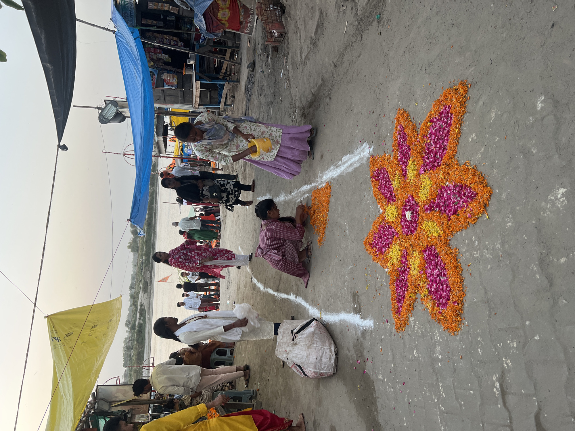 In Vrindavan, India, a group of people are creating a floral design on the ground. The design is a large, elaborate flower shape made of orange, pink, and yellow flower petals. Two individuals are directly involved in the creation: one woman is pouring something from a yellow container, presumably to outline the design or help the petals adhere, while another person is kneeling and carefully placing petals within the outline. A white powder line marks the boundary of the design area. Other people are standing nearby, observing the process, or are passing by. Overhead, there are colorful tarpaulins or tents. The scene appears to be taking place in a public area, possibly near a river or body of water, with simple structures and shops visible in the background.