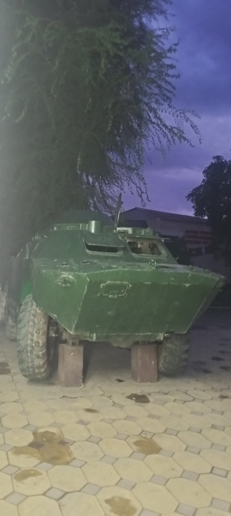 The image depicts a green armored personnel carrier (APC) parked on a paved area, possibly a courtyard or a parking lot, in Sokuluk, Kyrgyzstan. The APC is raised on wooden blocks, suggesting it might be undergoing maintenance or is on display. The vehicle has large, rugged tires and a distinctive angled hull. In the background, there are trees with dense foliage and a building with a visible roofline. The sky is a deep, twilight blue, indicating the photo was taken at dusk or dawn. The paving stones are hexagonal with darker grout lines. There are some dark stains on the paving stones, the origin of which is unclear. No people are visible in the image.