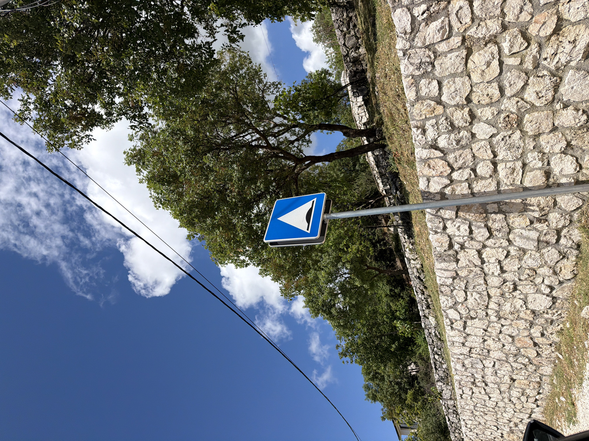The image captures a bright, sunny daytime scene in what appears to be a rural or semi-rural setting, specifically Bratešići, Montenegro. The sky, a vibrant blue, dominates the upper and left portions of the frame, adorned with scattered, fluffy white clouds.
On the right side of the image, a robust stone wall constructed from irregularly shaped, light-colored rocks stands prominently. Patches of green grass are visible along the base and top of this wall. To the left and top, dense green foliage from various trees creates a natural canopy, with sunlight filtering through the leaves. One large tree with visible branches is particularly central on the left side of the frame.
A blue square road sign with a white border is positioned towards the center-right, mounted on a silver metallic pole. The sign features a white arrow, depicted as two chevrons, pointing diagonally upwards and to the left, indicating a directional warning or instruction, likely for a sharp bend or obstacle.
Two dark utility lines or cables stretch diagonally across the sky from the lower left to the upper right, adding a man-made element to the natural landscape. No people or activities are visible. The scene is static, highlighting the natural beauty and infrastructural elements of the location under clear, bright weather conditions. There is no discernible text on the sign, only the graphic symbol.