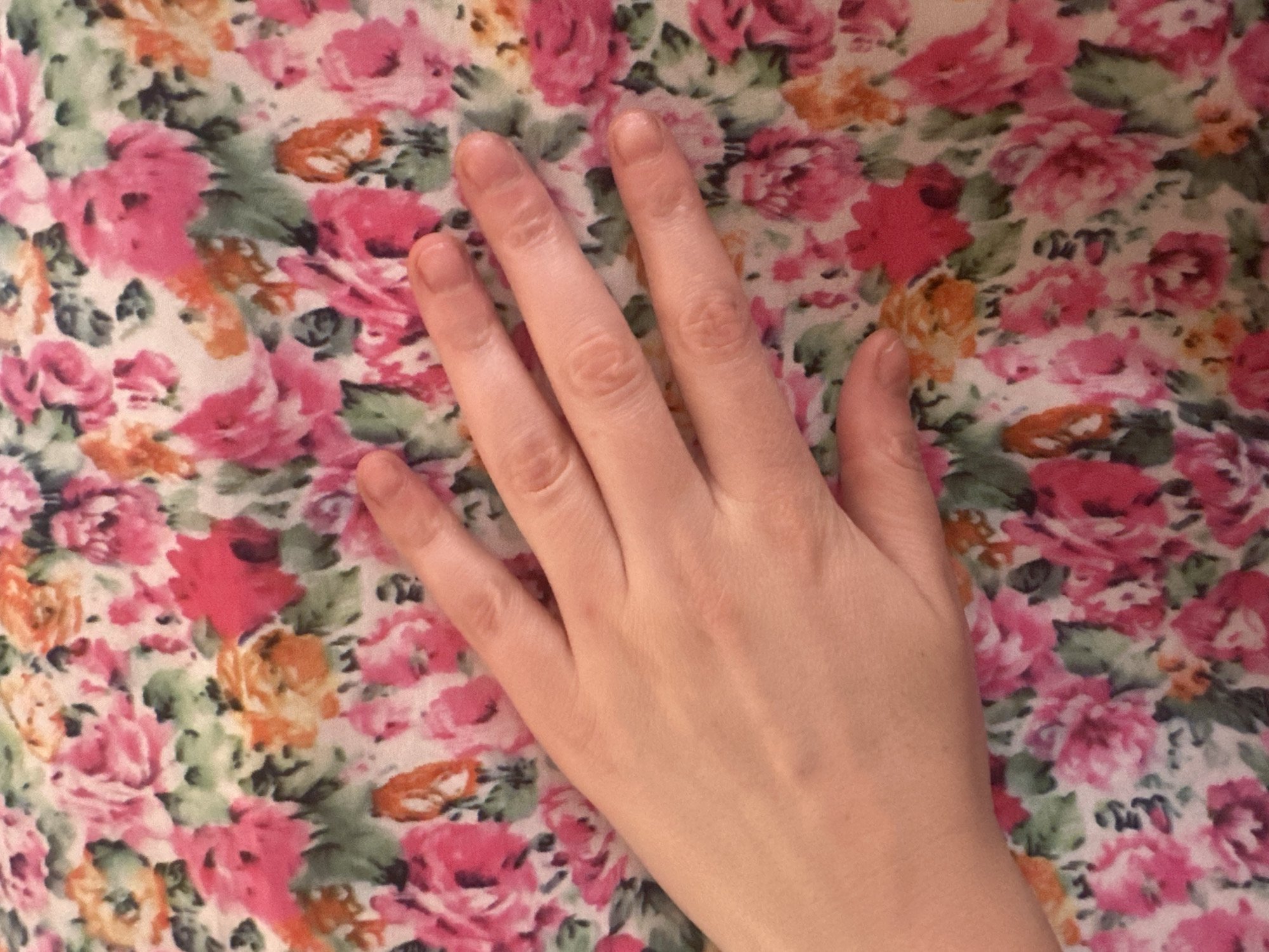 An overhead medium shot shows a person's left hand resting on a floral patterned fabric. The fabric features a dense arrangement of pink, fuchsia, and orange roses with green leaves. The hand, with its fingers slightly splayed, is positioned diagonally across the frame, with the palm and fingers in focus against the textured background. The skin tone of the hand is fair, and the fingernails are bare. The lighting is soft, casting no harsh shadows. There is no discernible urban environment present in the image; it is a close-up of a hand and fabric.